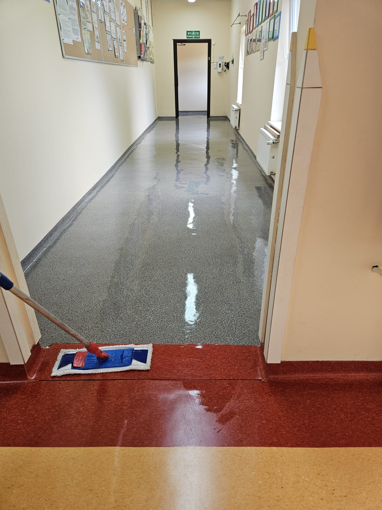 Mop z niebieską nakładką czyszczący błyszczącą posadzkę w korytarzu z tablicą ogłoszeń i oknami.