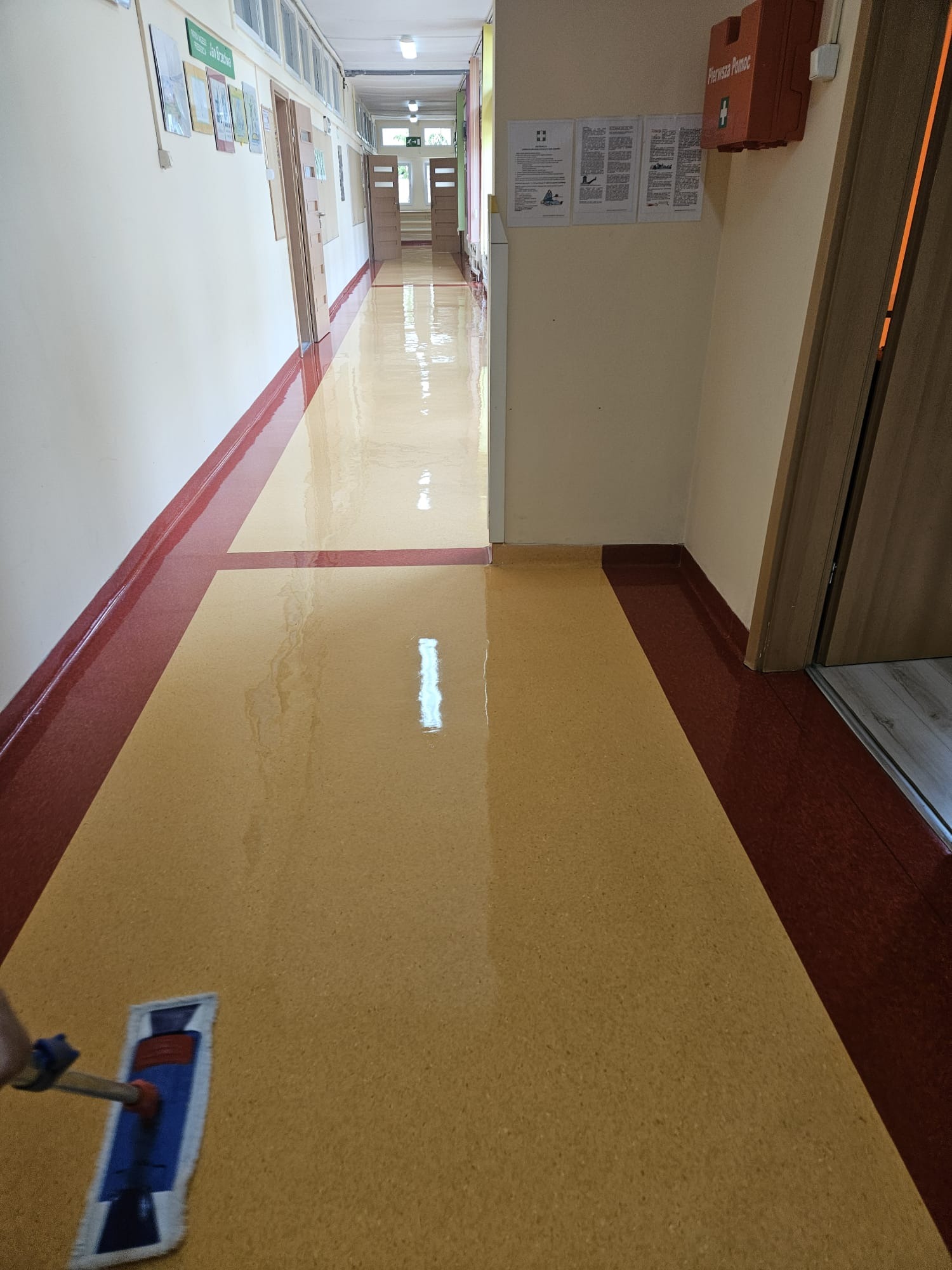 Mop z niebieską nakładką czyszczącą na długim kiju czyści błyszczącą podłogę w korytarzu szkolnym z czerwonymi listwami przypodłogowymi, w tle tablice informacyjne i apteczka pierwszej pomocy.