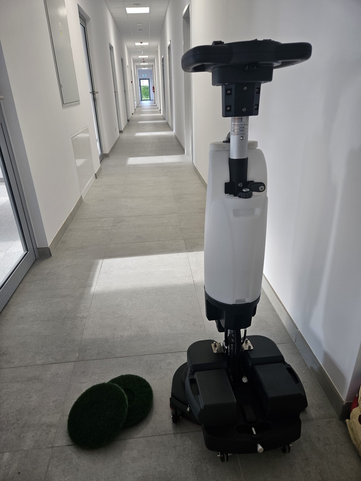 Automat szorująco-zbierający do podłóg na tle długiego, pustego korytarza z szarymi płytkami i białymi ścianami. Dwie zielone podkładki do szorowania leżą obok maszyny.