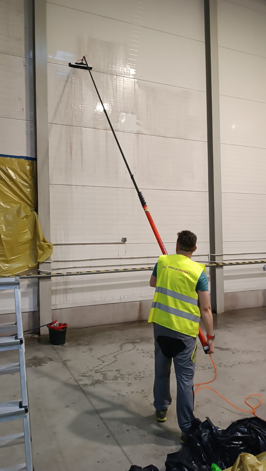 Pracownik w żółtej kamizelce odblaskowej czyści wysoką, białą ścianę hali przemysłowej za pomocą mopa na długim kiju z czerwonym uchwytem. Na ścianie widoczne ślady wody.