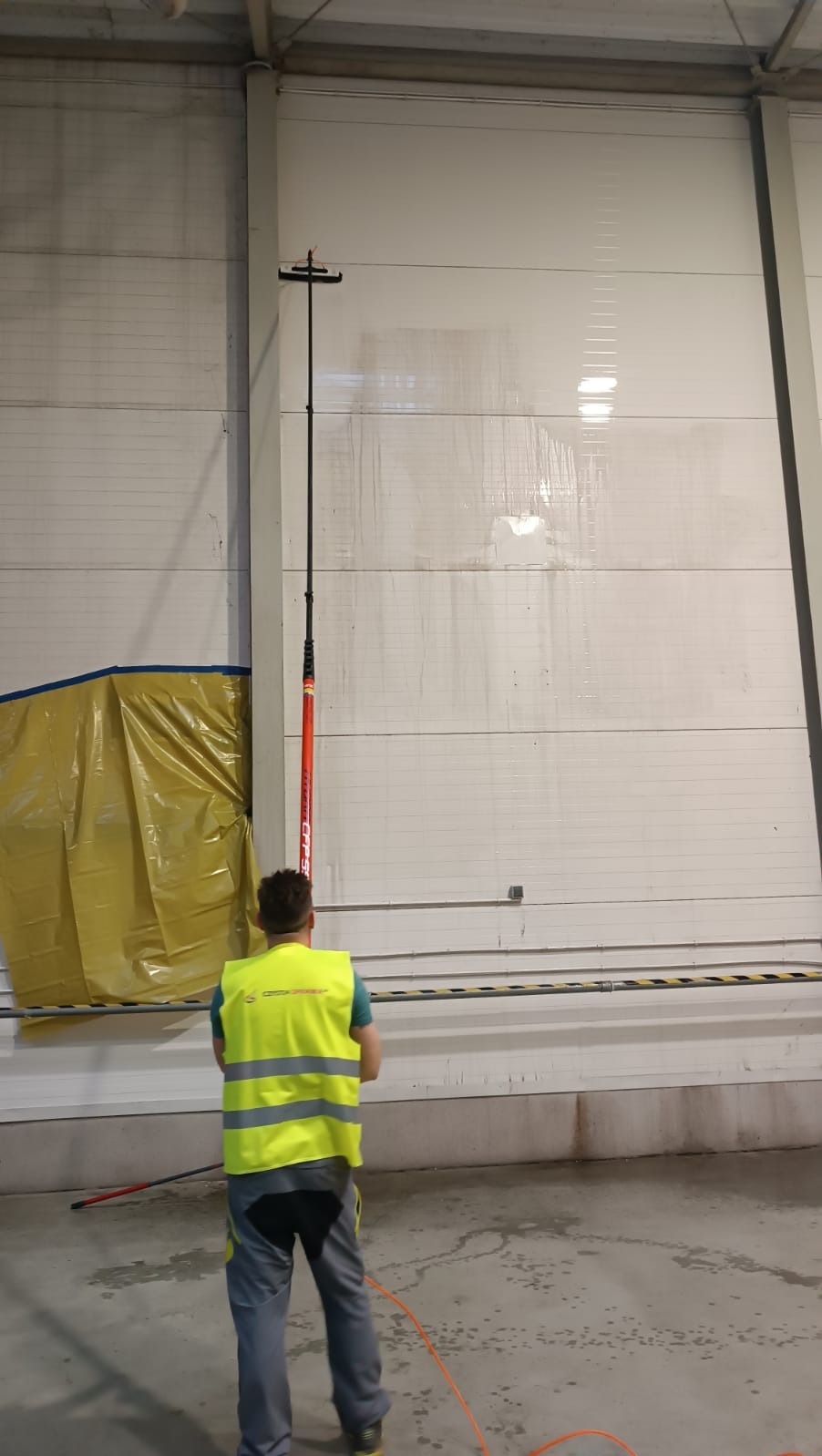 Pracownik w kamizelce odblaskowej czyści wysoką bramę przemysłową za pomocą teleskopowego kija z myjką, widoczne ślady czyszczenia na panelach bramy.