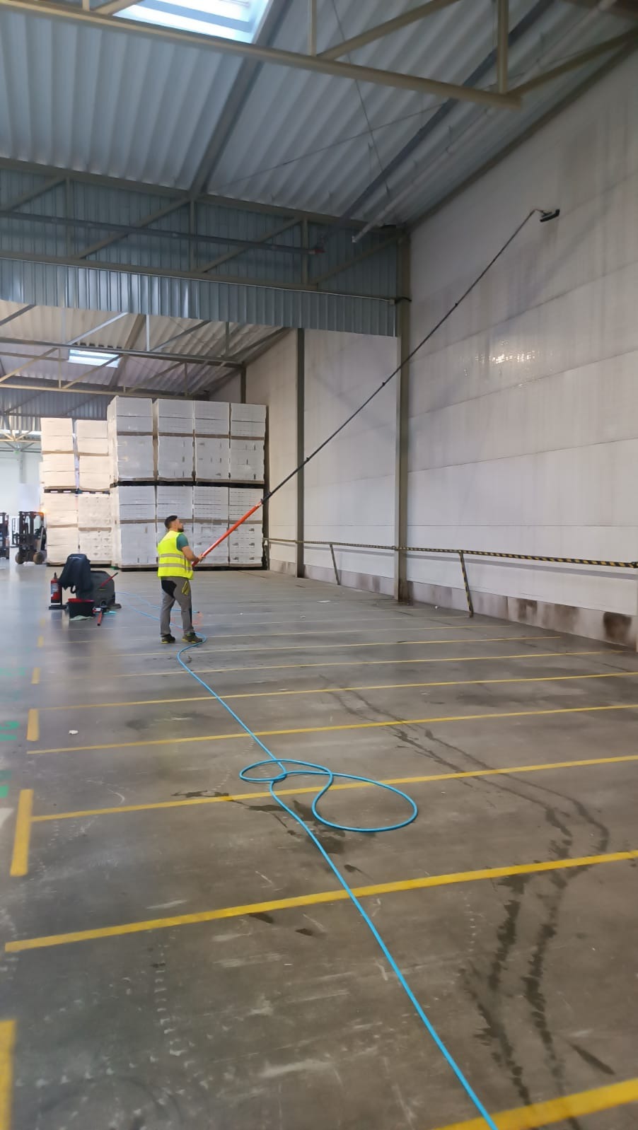 Pracownik w kamizelce odblaskowej czyści wysoką ścianę hali magazynowej za pomocą lancy teleskopowej z zamontowaną szczotką, niebieski wąż leży na podłodze z żółtymi liniami.