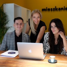 MIESZKANIE.COM SPÓŁKA Z OGRANICZONĄ ODPOWIEDZIALNOŚCIĄ - Zespół trzech agentów nieruchomości uśmiecha się za laptopem na drewnianym biurku, z logo 'mieszkanie.com' na żółtej ścianie w tle.