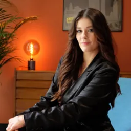 Kobieta w czarnym garniturze siedzi na turkusowym fotelu w biurze z pomarańczowymi ścianami, na których wisi plakat z motywem miasta, a obok stoi lampka w stylu retro.