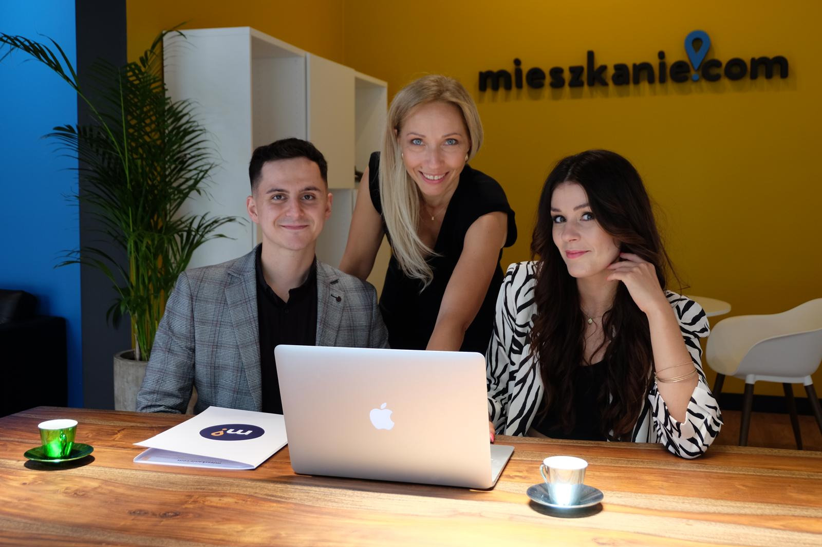 Zespół trzech agentów nieruchomości uśmiecha się za laptopem na drewnianym biurku, z logo 'mieszkanie.com' na żółtej ścianie w tle.