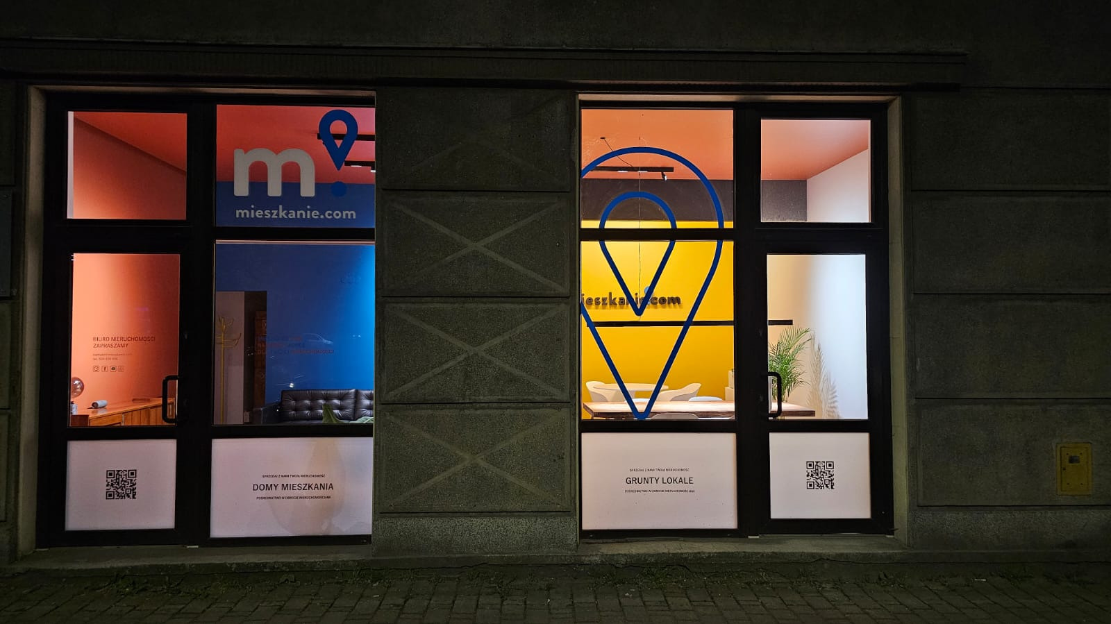 Dwa okna wystawowe agencji nieruchomości 'mieszkanie.com' z naklejonym logo i informacjami, oświetlone różnymi kolorami wewnątrz.