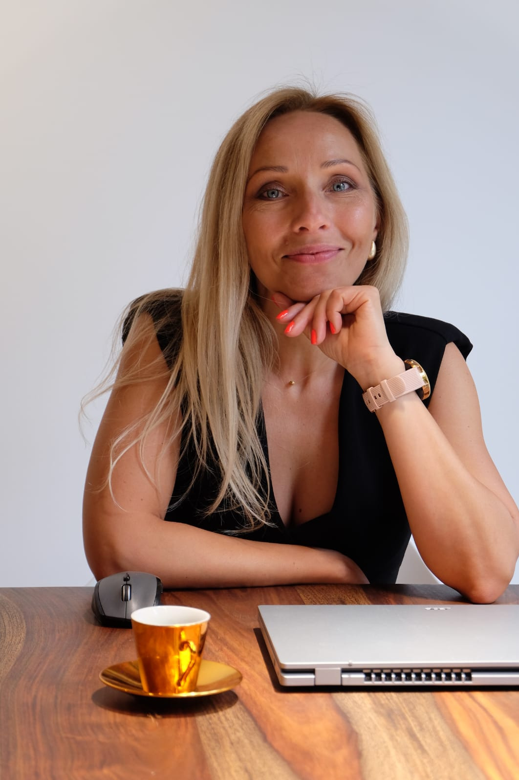 Uśmiechnięta blondynka z różowym smartwatchem, oparta o dłoń, siedzi przy biurku z laptopem i złotą filiżanką.