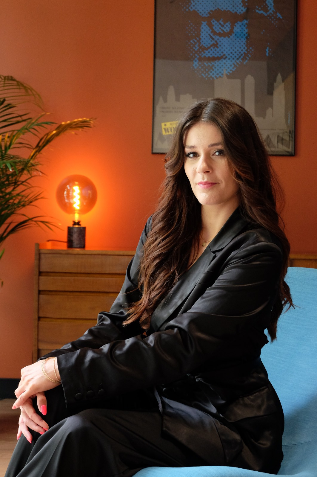 Kobieta w czarnym garniturze siedzi na turkusowym fotelu w biurze z pomarańczowymi ścianami, na których wisi plakat z motywem miasta, a obok stoi lampka w stylu retro.