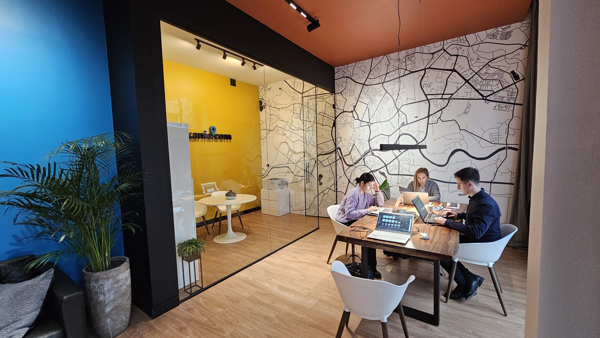 Nowoczesne biuro agencji nieruchomości z trzema osobami pracującymi przy laptopach, mapa miasta na ścianie, żółty pokój konferencyjny za szklaną ścianą.