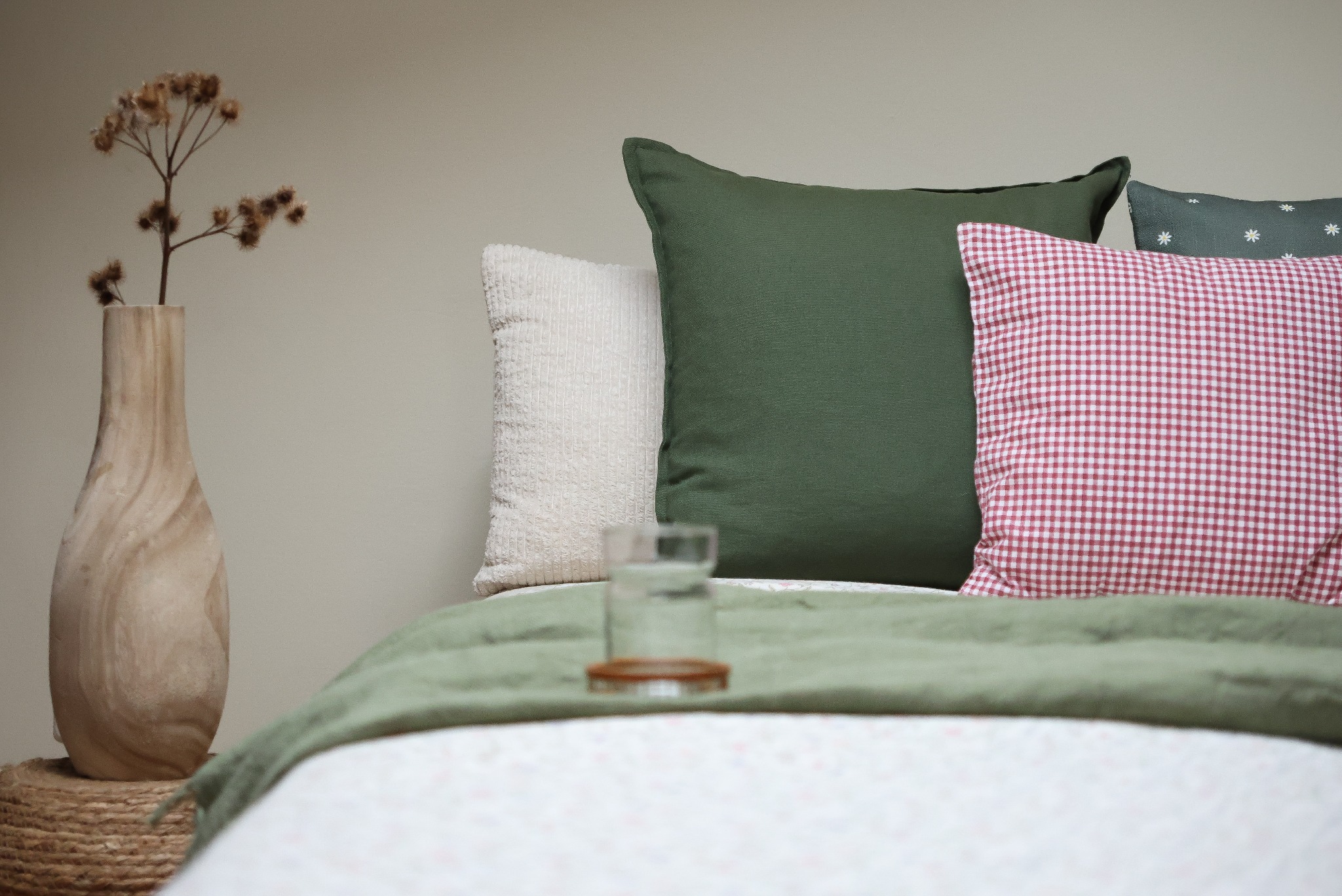 Dekoracyjna kompozycja na łóżku: wazon z suchymi kwiatami na plecionym stoliku, obok szklanka wody, a w tle poduszki w odcieniach bieli, zieleni i czerwono-białej kraty.