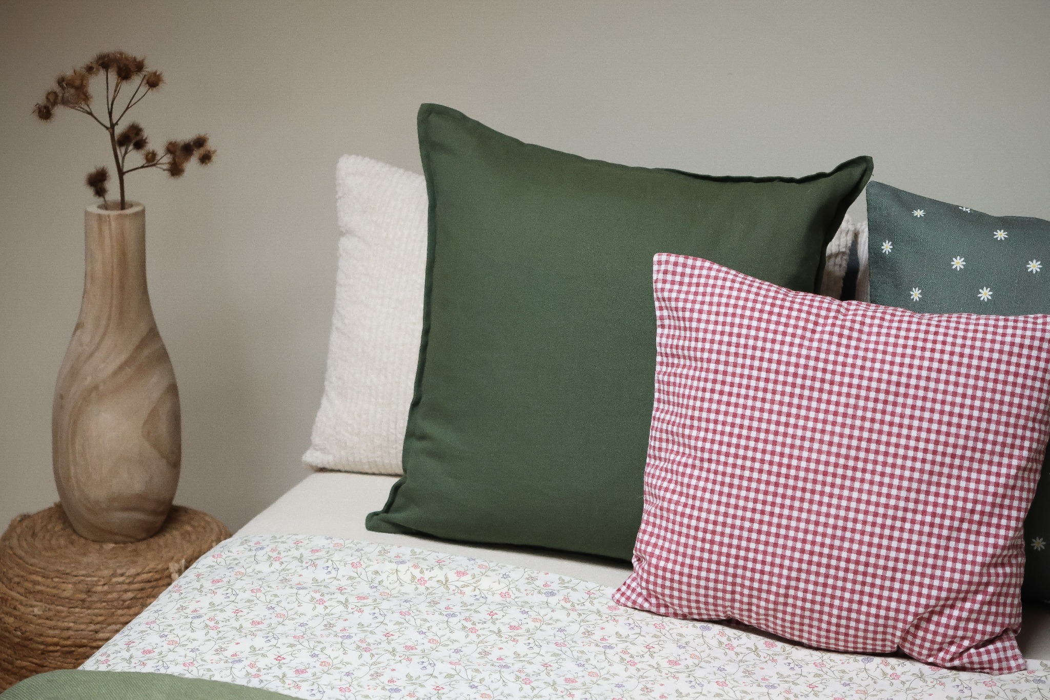Dekoracyjne poduszki w odcieniach zieleni i czerwieni w kratę na łóżku z pościelą w drobne kwiatki, obok drewniany wazon z suchymi gałązkami.
