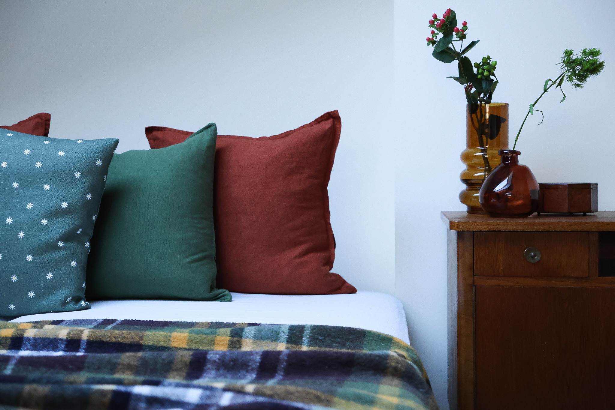 Aranżacja sypialni z trzema poduszkami w odcieniach niebieskiego, zielonego i ceglastego, pled w kratę na łóżku oraz drewniana szafka nocna z brązowymi wazonami i szkatułką.