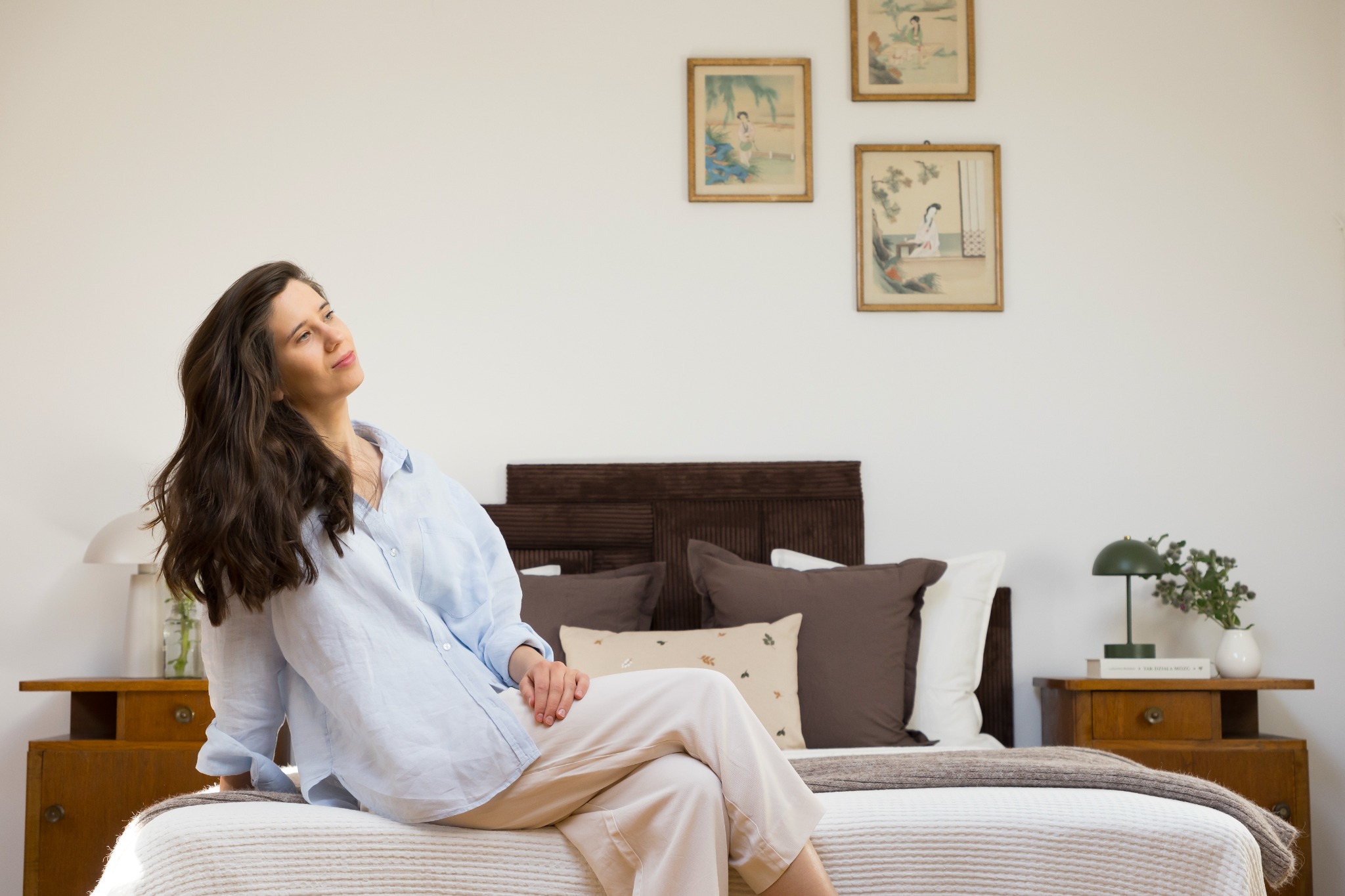Jasna sypialnia z brunetką w błękitnej koszuli siedzącą na łóżku. Nad łóżkiem wiszą trzy obrazki w złotych ramkach. Na szafce nocnej lampka i wazon z kwiatami.