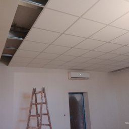 Anatolii Kardynal - Biały sufit kasetonowy z widoczną konstrukcją nośną w trakcie montażu, drabina oparta o ścianę, klimatyzator na ścianie. Widok na wnętrze w trakcie prac wykończeniowych.