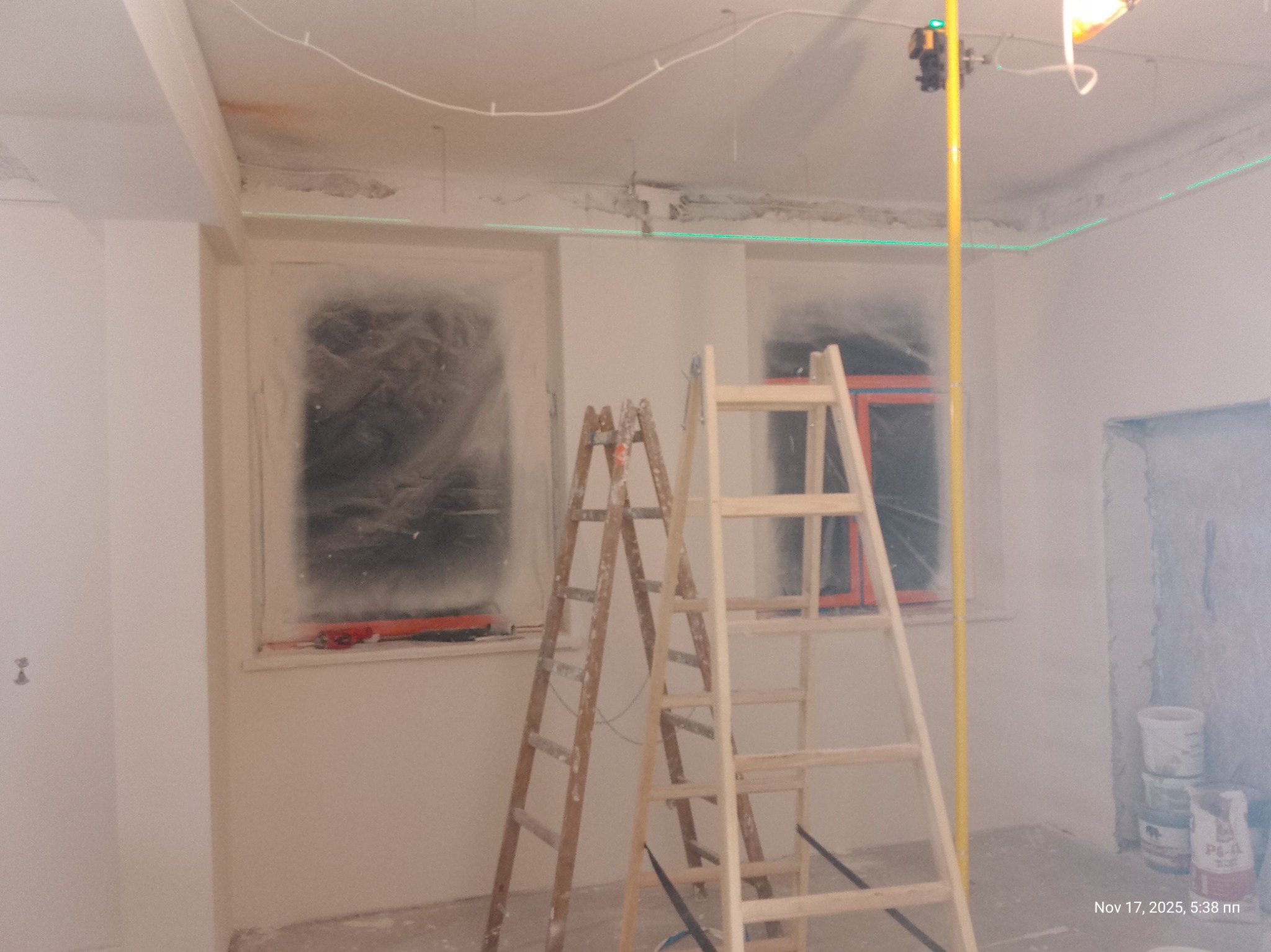 Pokój w trakcie remontu: okna zabezpieczone folią, dwie drabiny, laser krzyżowy wyznacza poziom. Widoczne ślady po starych listwach sufitowych.