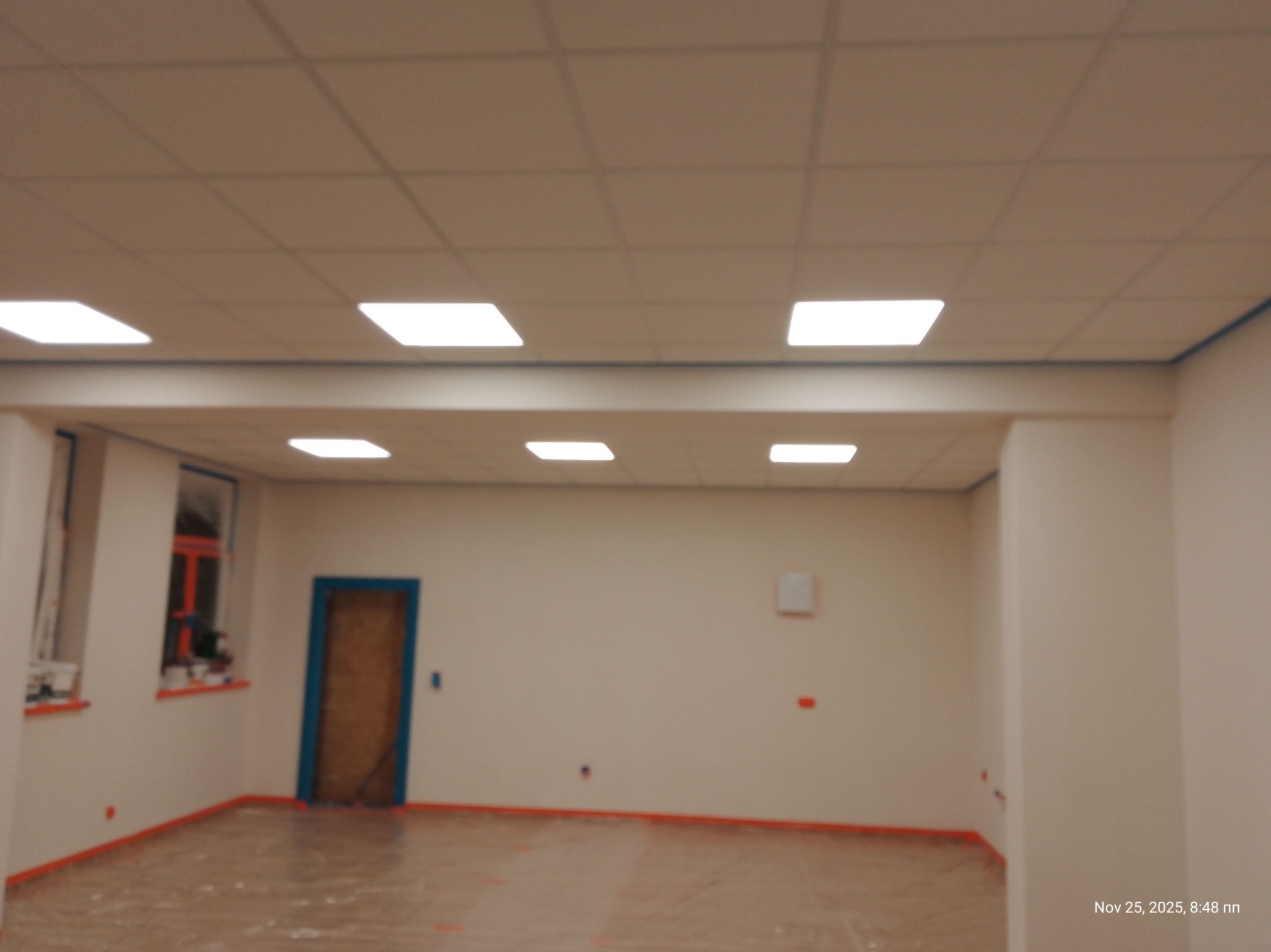 Pokój po malowaniu ścian na biało, zabezpieczony folią i taśmą malarską. Widoczne okno i drzwi z niebieskimi ramami, oraz sufit z kwadratowymi lampami.