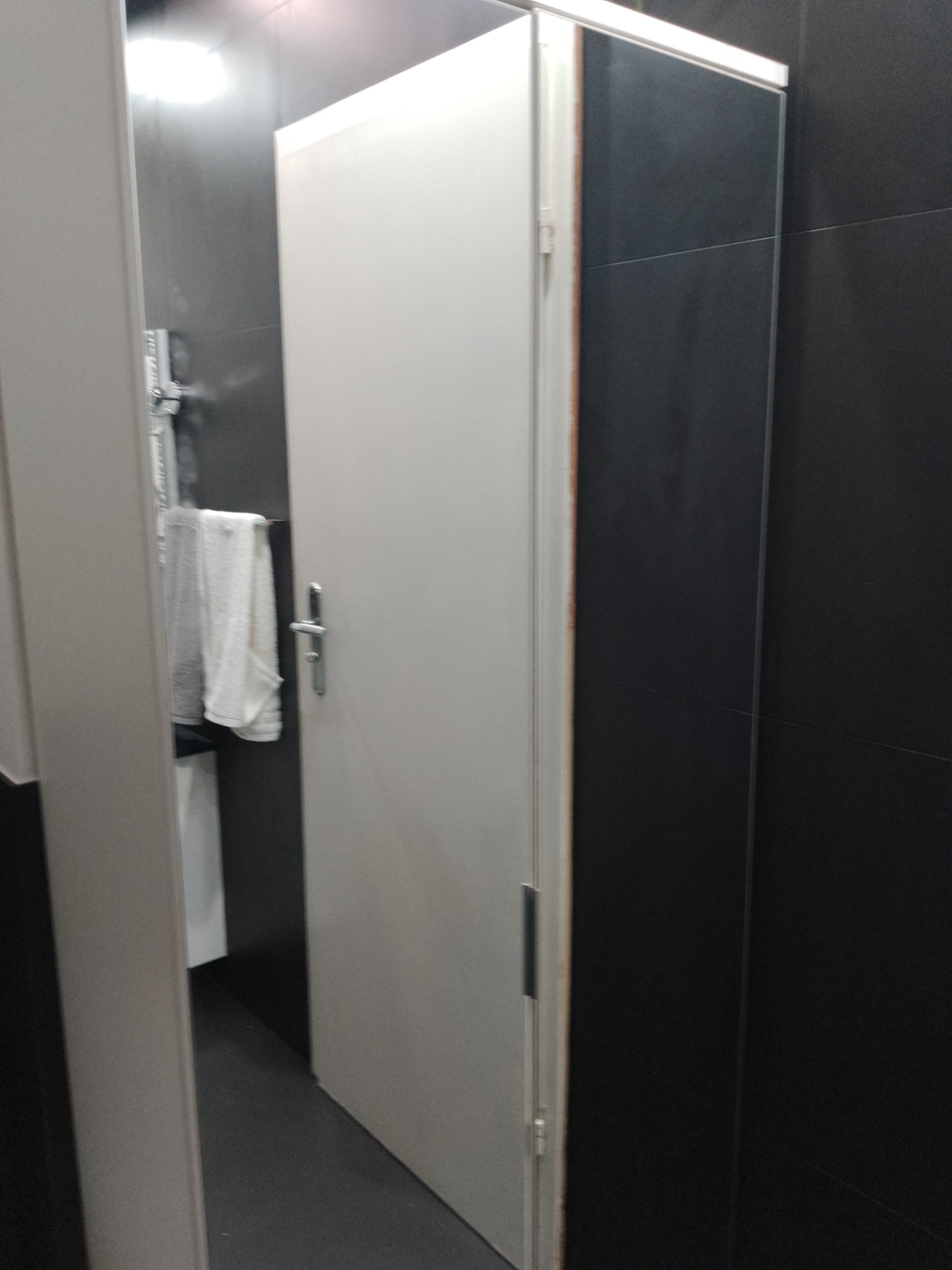 Nowoczesna kabina prysznicowa z białą ramą i drzwiami, czarne płytki na ścianach, widoczny fragment baterii i ręcznika wewnątrz kabiny.