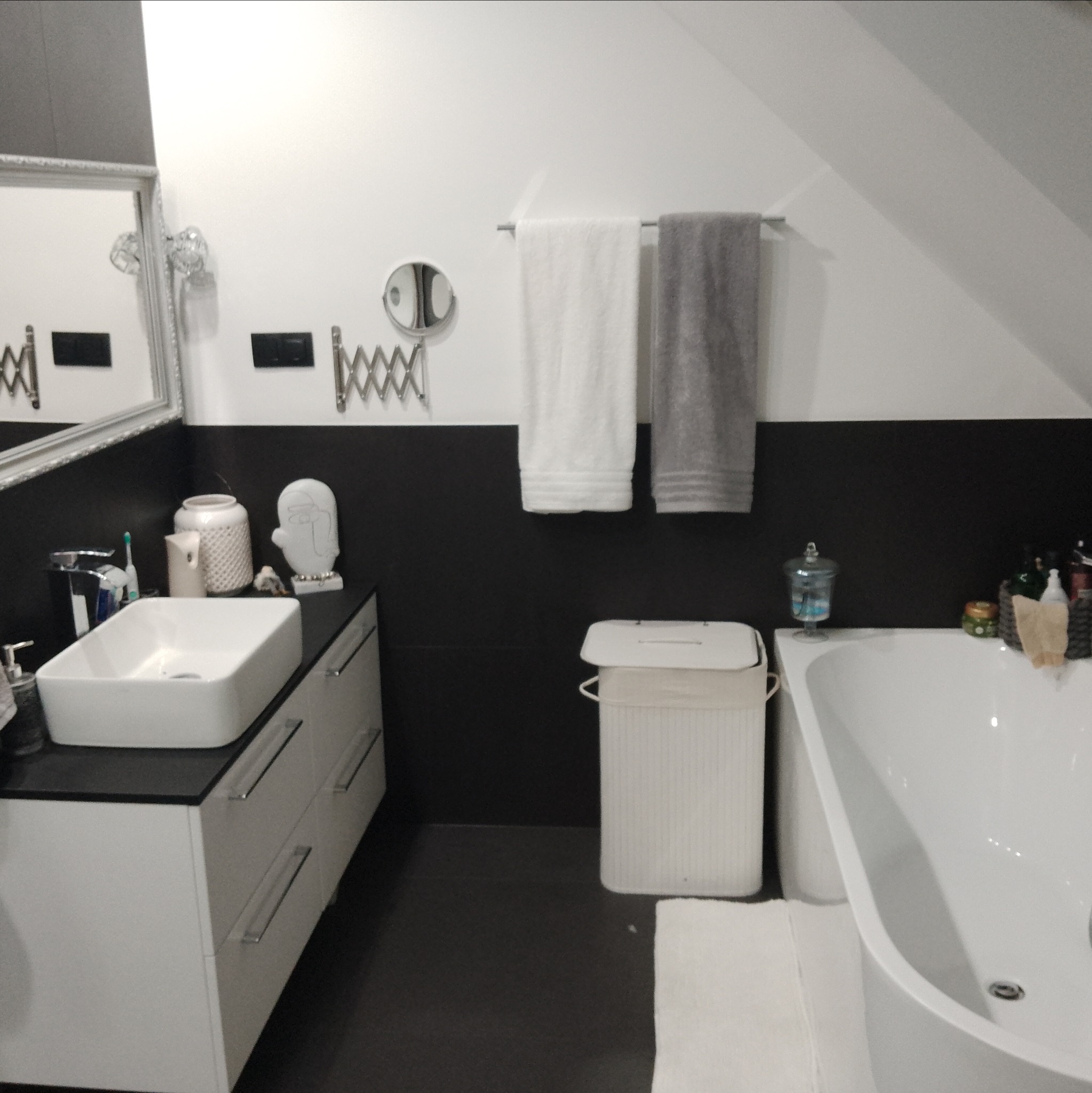 Nowoczesna łazienka na poddaszu z białą umywalką nablatową, czarną ścianą do połowy wysokości i wiszącymi ręcznikami. Minimalistyczny design.