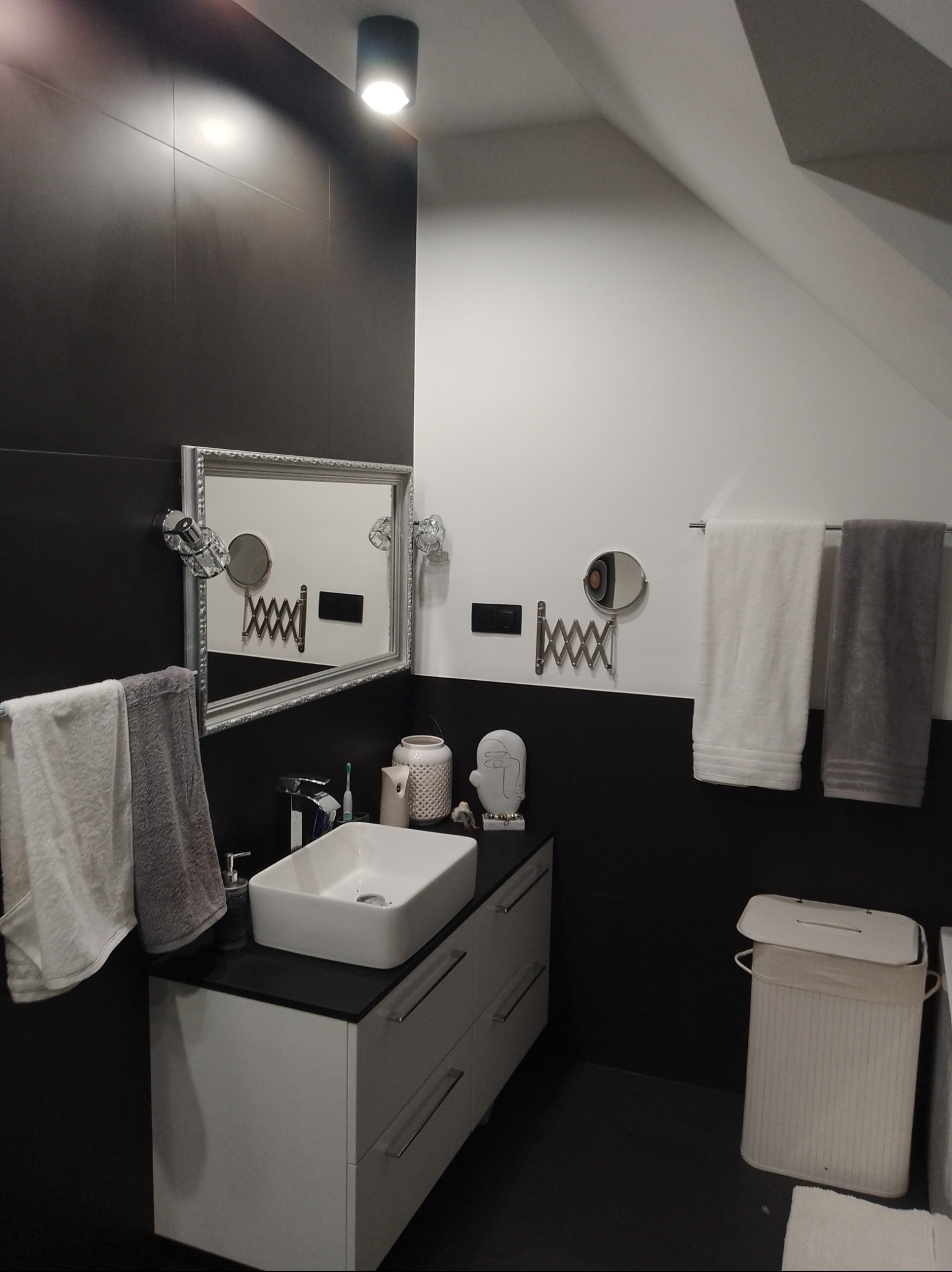 Nowoczesna łazienka w czerni i bieli: prostokątna umywalka nablatowa, lustro w ozdobnej ramie, czarne płytki do połowy ściany, białe ręczniki i dekoracje.