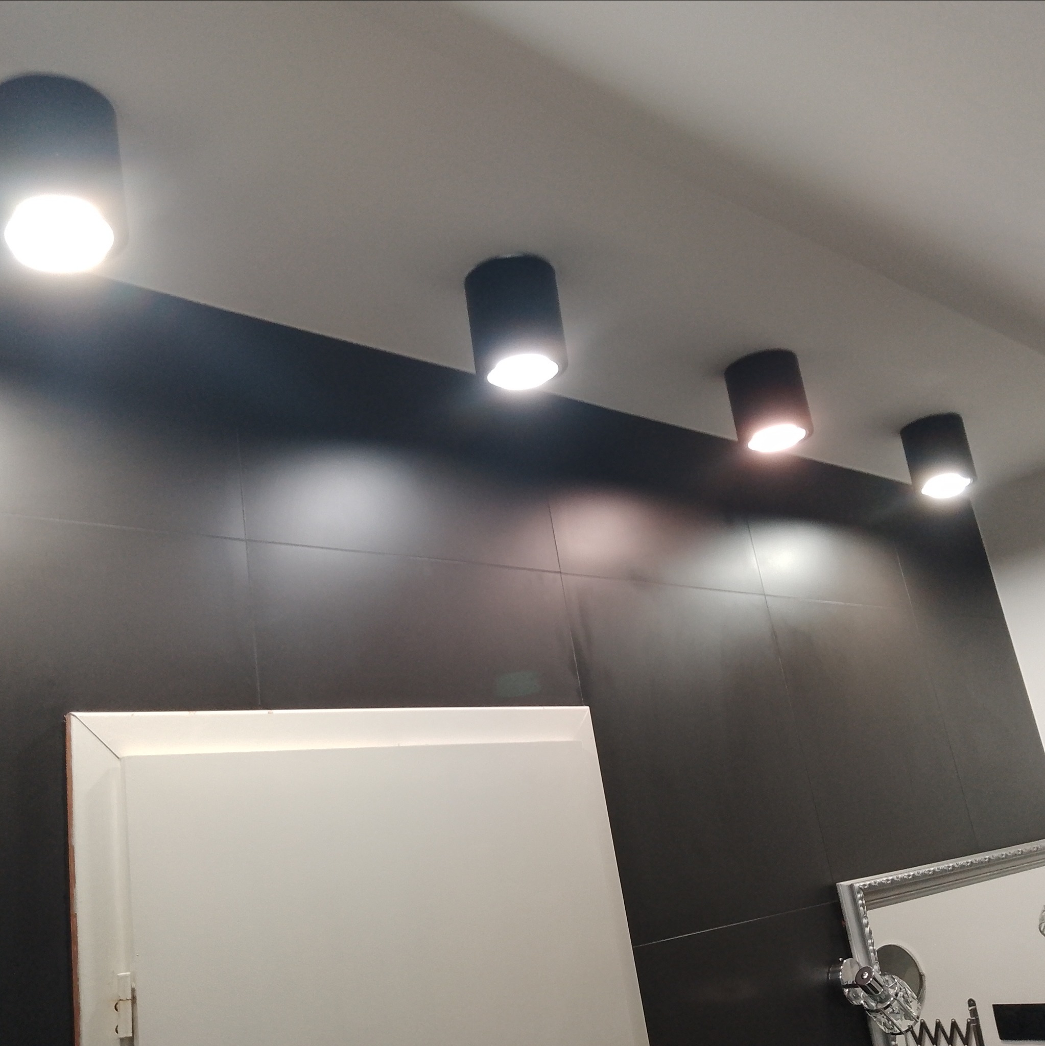 Czarne, cylindryczne lampy sufitowe LED o ciepłej barwie światła, zamontowane na tle ciemnej, błyszczącej ściany z płytek. Widoczny fragment białej szafki i dekoracyjne elementy.