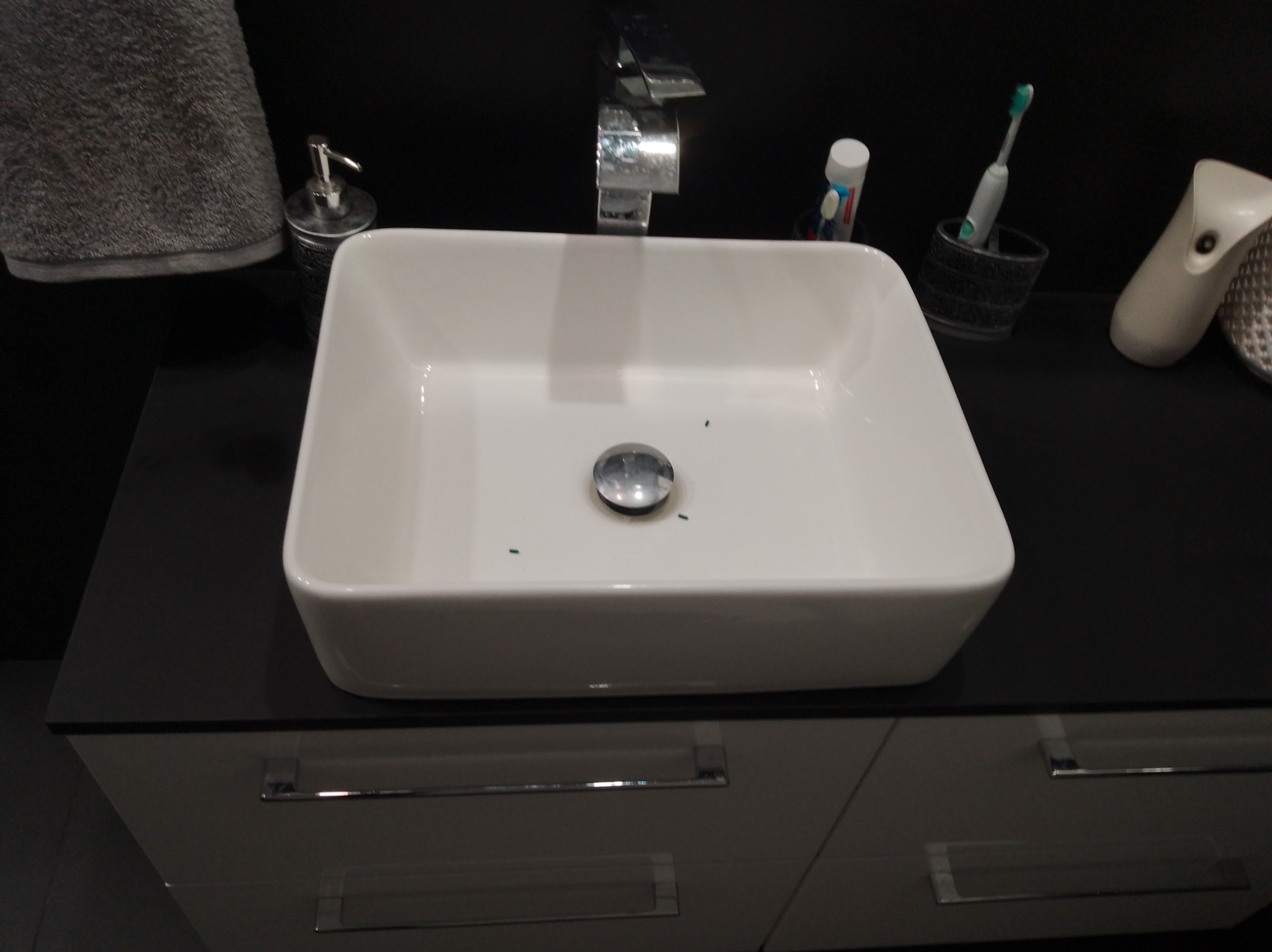 Biała, prostokątna umywalka nablatowa z chromowaną baterią na czarnym blacie szafki łazienkowej z szufladami. Widoczny dozownik mydła i akcesoria łazienkowe.