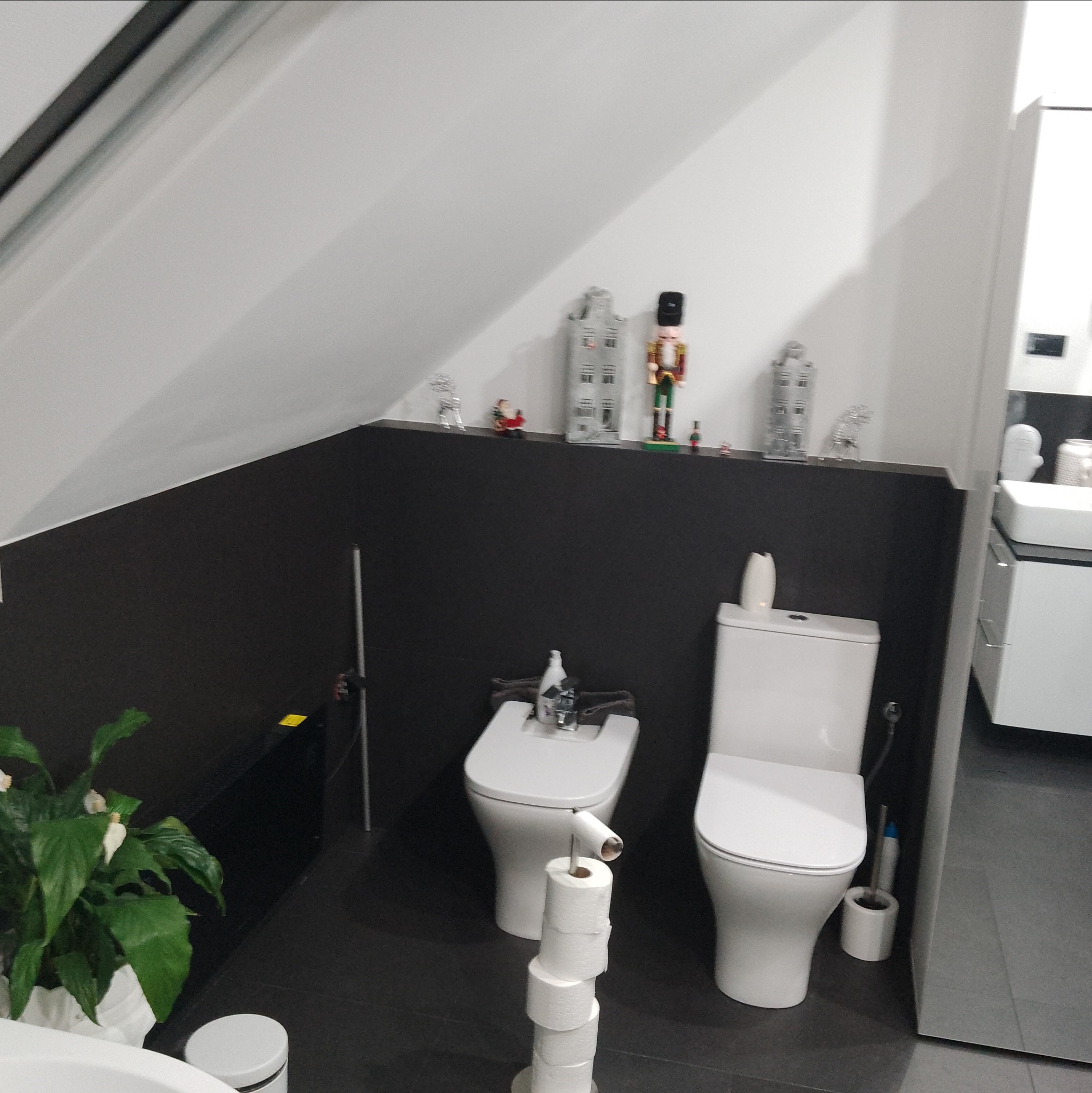 Nowoczesna łazienka na poddaszu z czarną ścianą, białymi sanitariatami i dekoracjami na półce. Minimalistyczny design z rośliną w rogu.