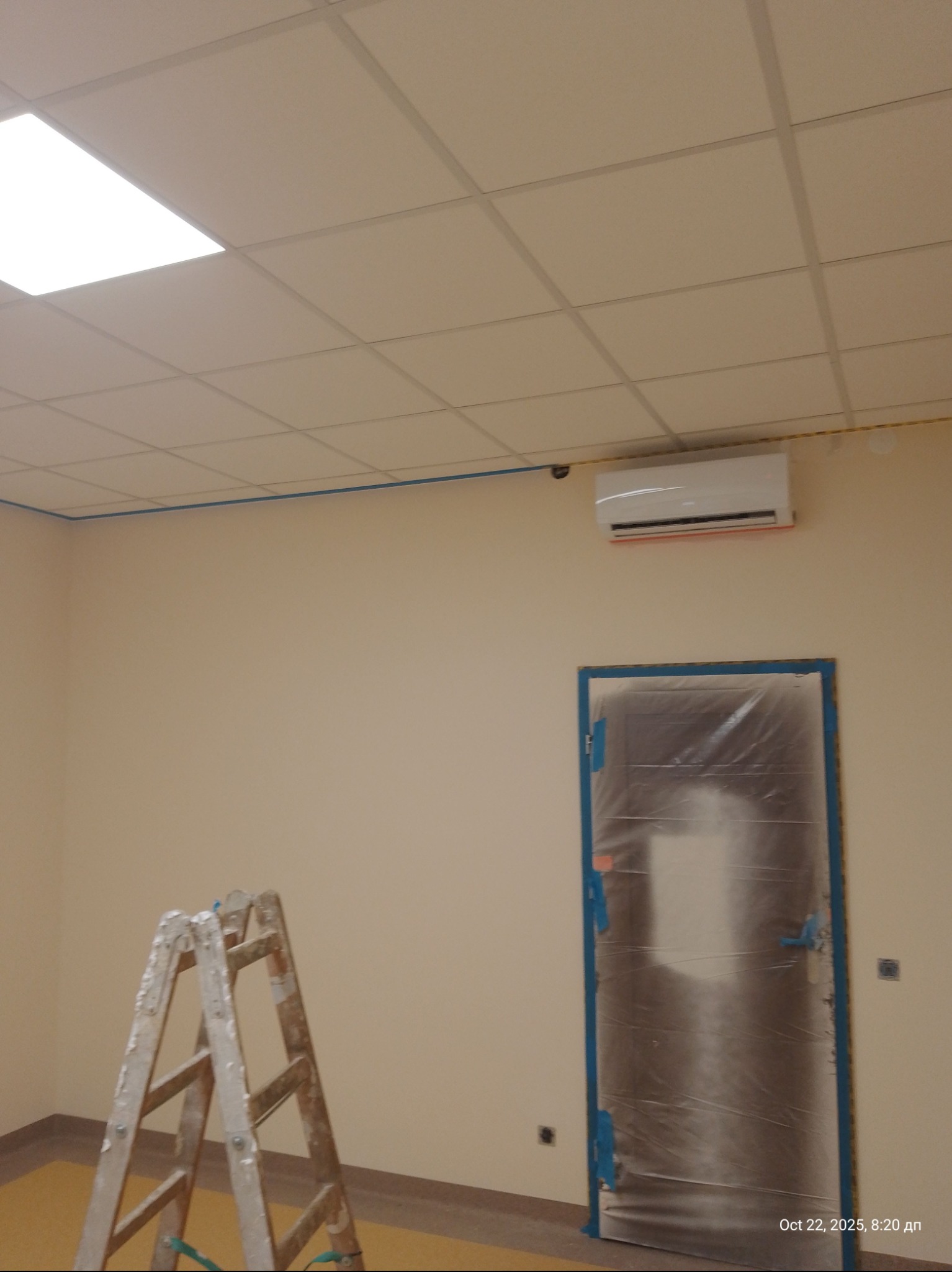 Pokój w trakcie malowania: drabina, zabezpieczone folią drzwi, klimatyzator na ścianie. Widoczne ślady taśmy malarskiej. Sufit kasetonowy z oświetleniem.