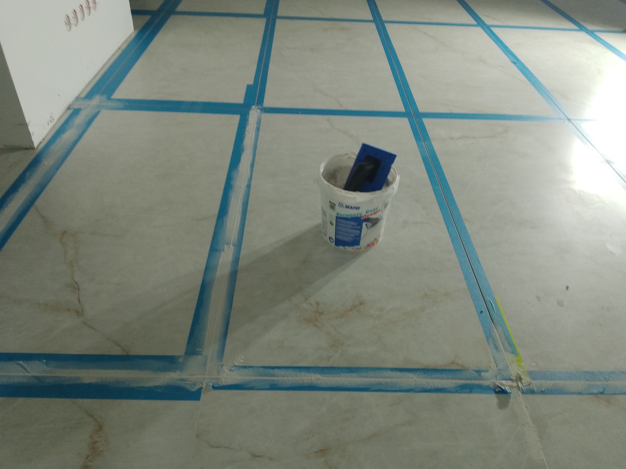 Świeżo ułożona, jasna podłoga z marmurowych płytek, zabezpieczona niebieską taśmą. Wiadro z zaprawą i szpachelka w centrum kadru. Widok z góry, ukończony etap prac glazurniczych.