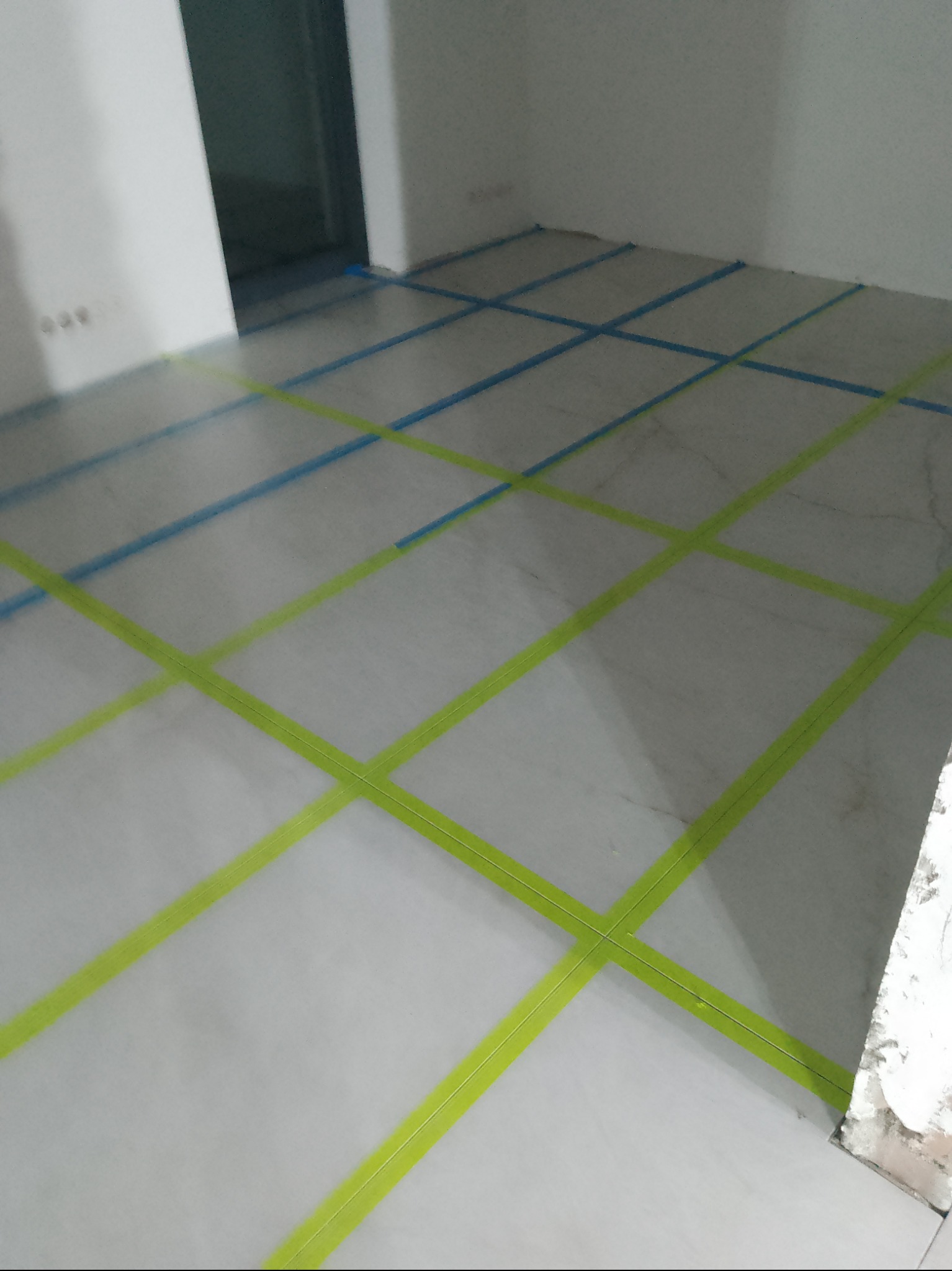 Przygotowanie podłoża pod płytki: podział powierzchni taśmą malarską w kolorze zielonym i niebieskim. Widok na podłogę w trakcie prac glazurniczych.