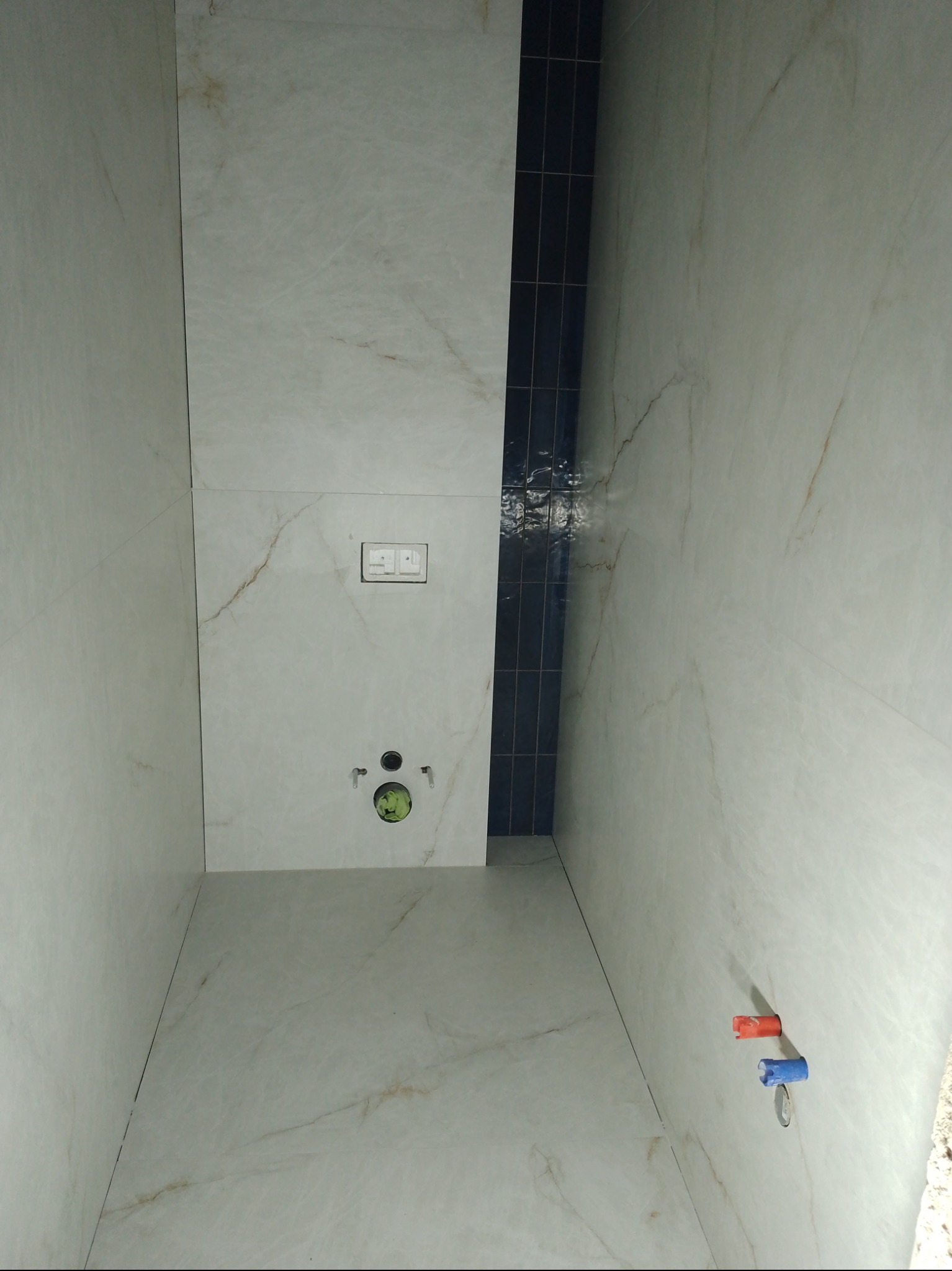 Wykończona łazienka z marmurowymi płytkami, pionowy pas ciemnych płytek, przyciski spłuczki i wystające rury. Widok z dołu, podkreślający geometrię i detale wykończenia.