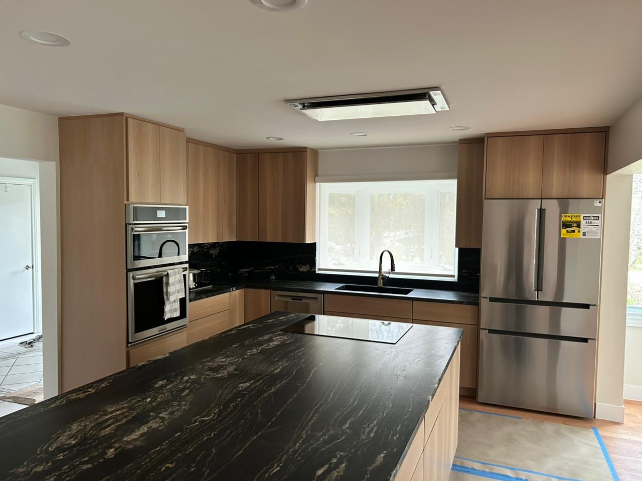 Nowoczesna kuchnia z drewnianymi szafkami, czarnym blatem i wyspą. Widoczny sprzęt AGD: piekarnik, lodówka. Okno z roletą w tle. Minimalistyczny design.