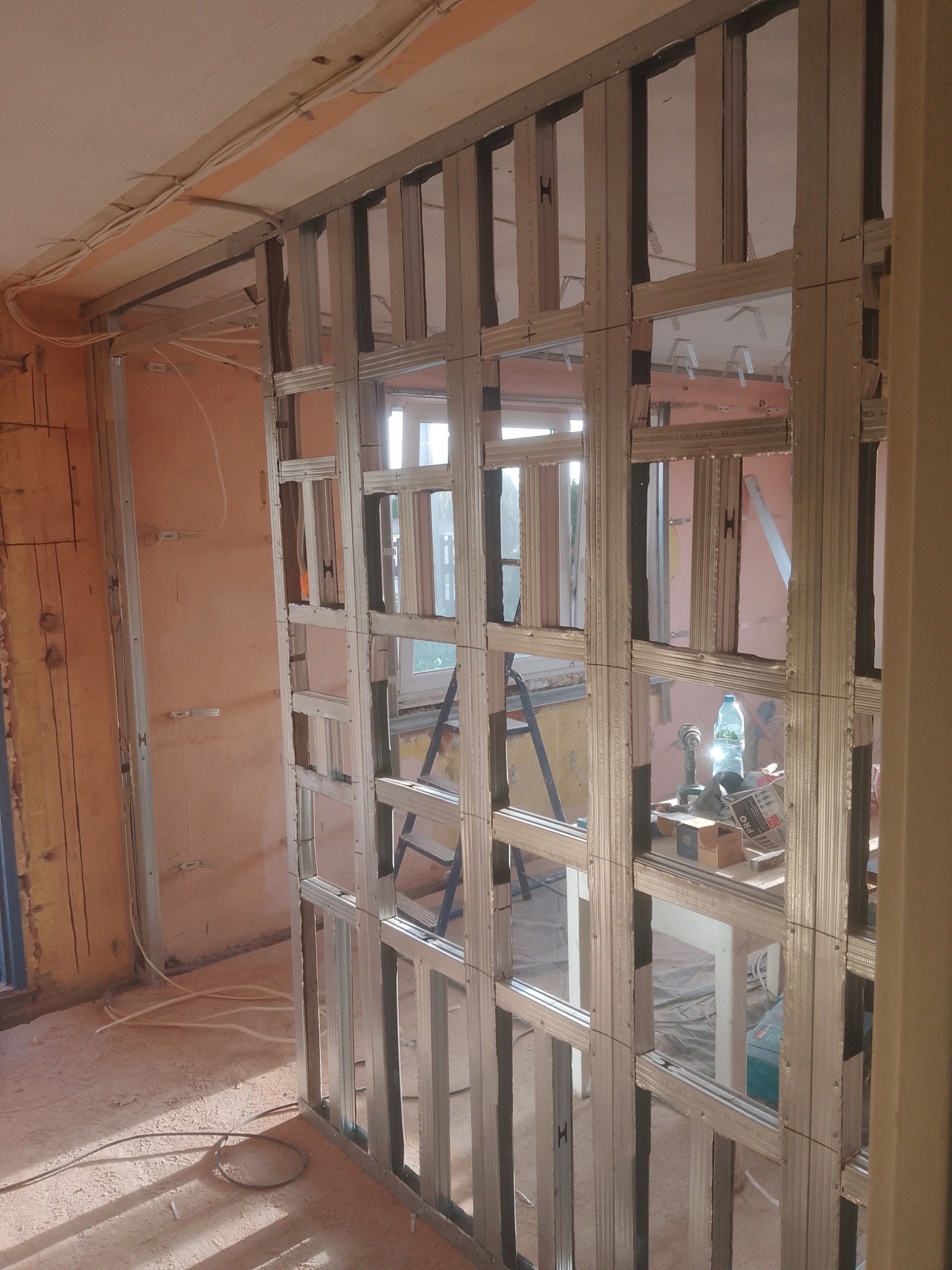 Metalowa konstrukcja ścianki działowej z profili, widoczne narzędzia i drabina w tle, surowy stan wnętrza w trakcie prac wykończeniowych.