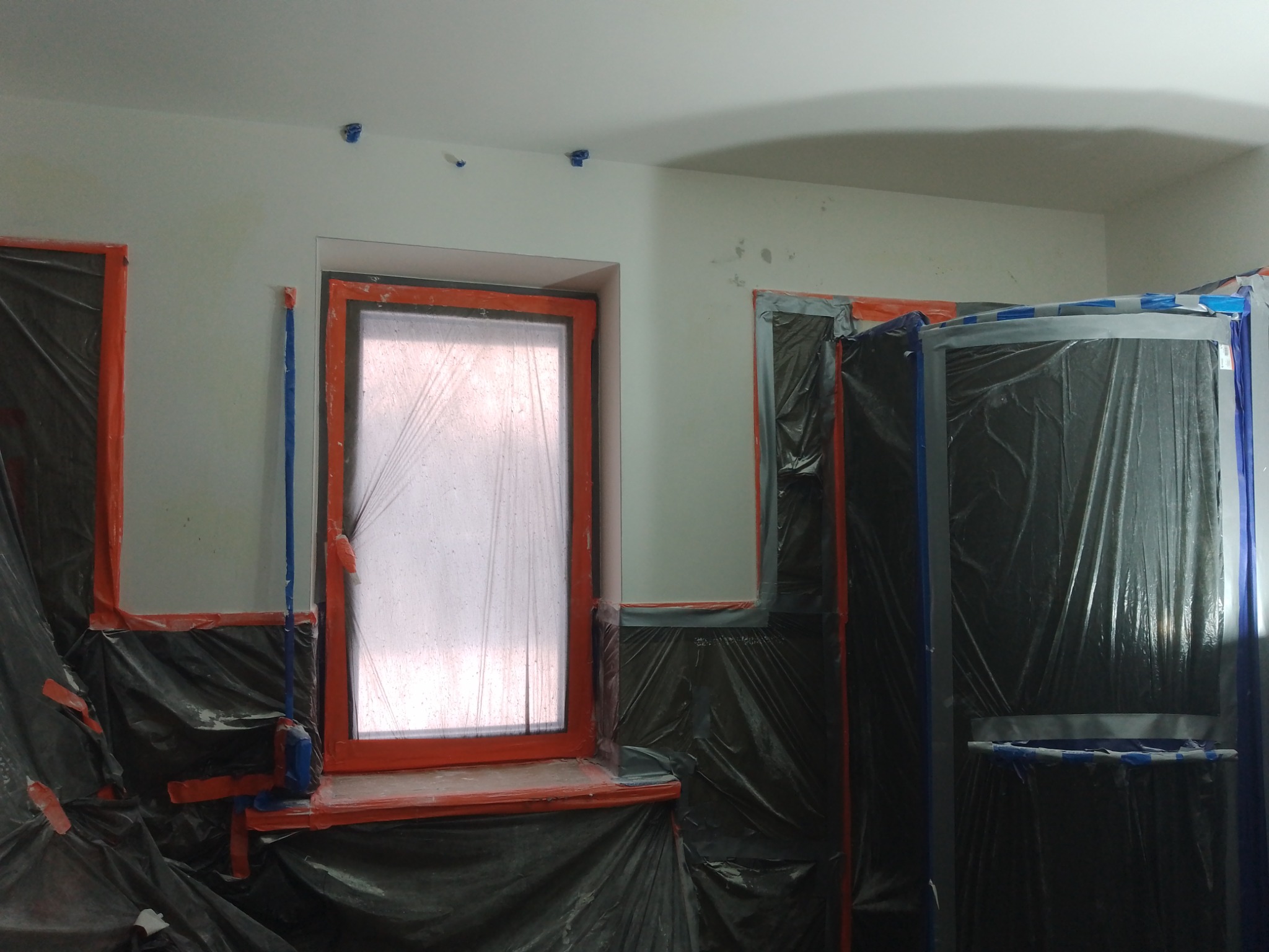 Pokój zabezpieczony folią malarską i taśmą przed malowaniem. Okna i meble osłonięte, przygotowane do prac wykończeniowych. Widoczne ślady po szpachlowaniu na suficie.