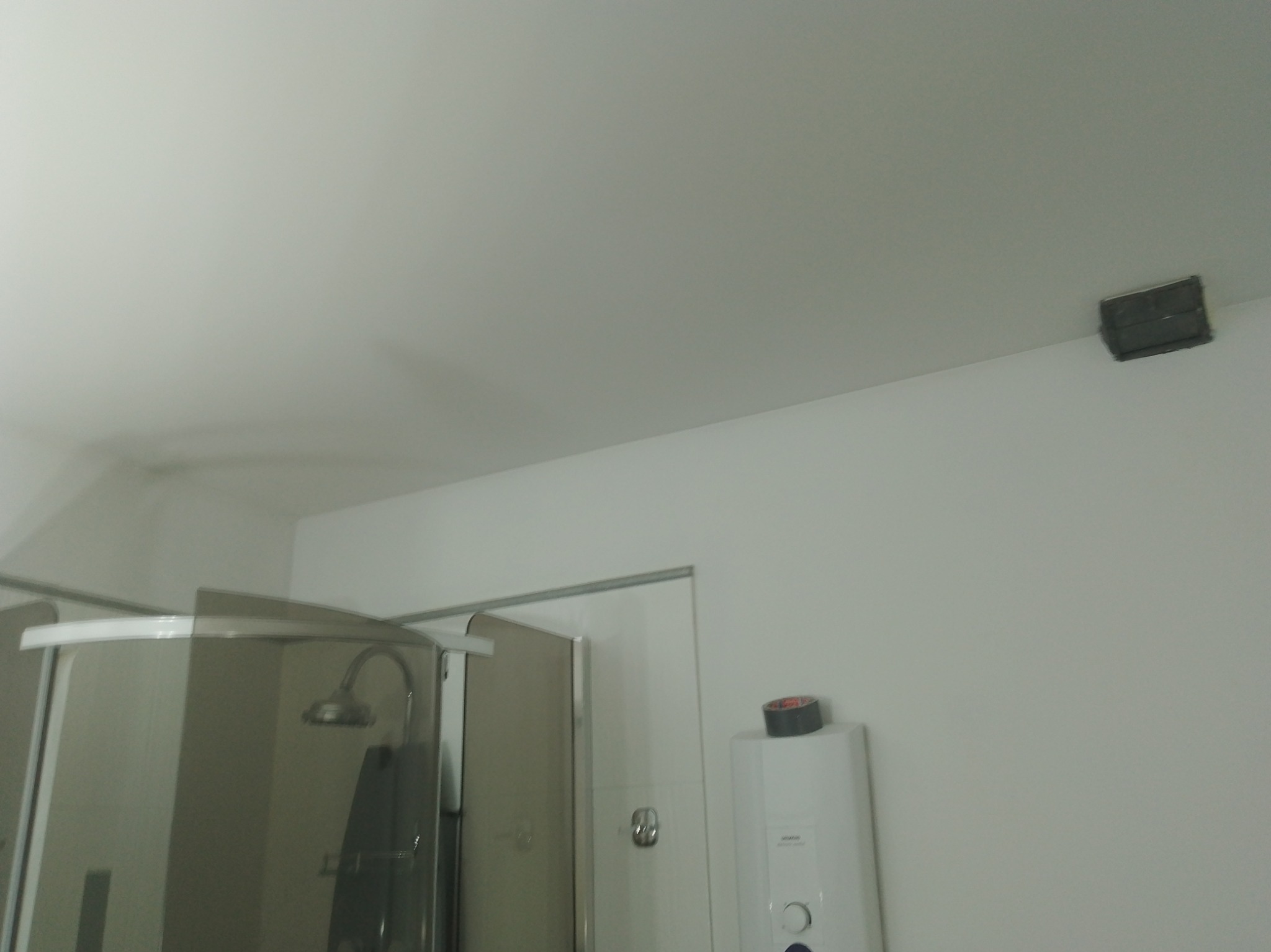 Biała łazienka z kabiną prysznicową i bojlerem, na którym leży taśma. Na suficie widoczna puszka elektryczna. Minimalistyczne wykończenie.