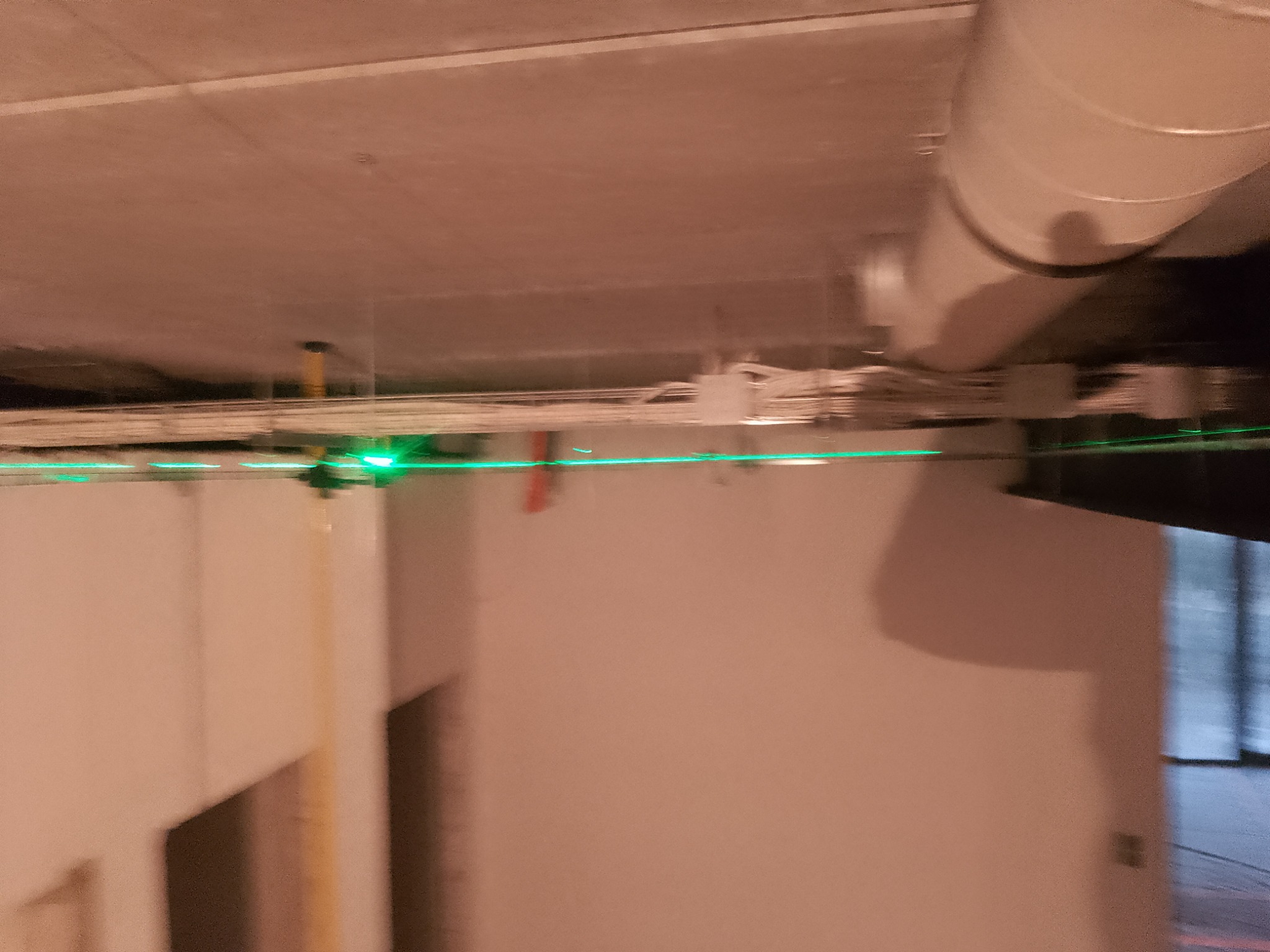 Zielona linia lasera wyznacza poziom na tle surowego sufitu z widocznymi kablami i rurami wentylacyjnymi. Przygotowanie do prac wykończeniowych.