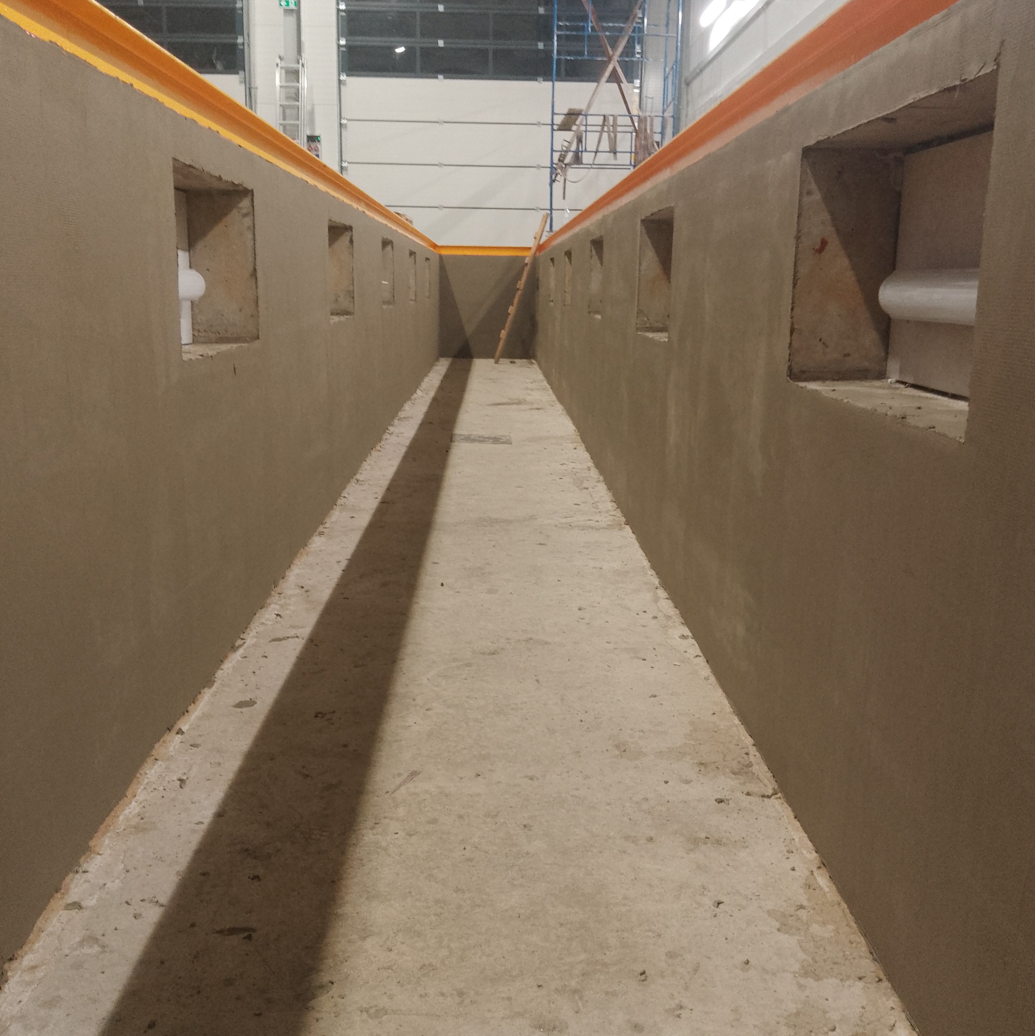 Wnętrze w trakcie szpachlowania ścian z oknami, widoczny surowy beton na podłodze i drabina w tle. Prace wykończeniowe w nowym budynku.