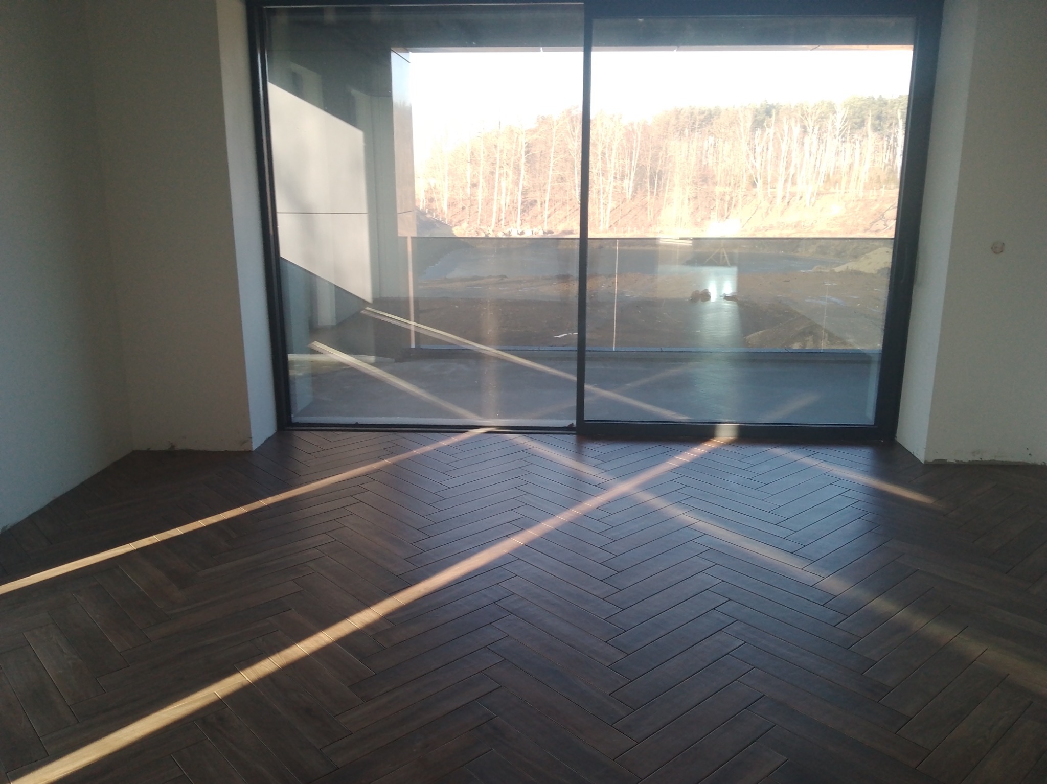 Ułożona, ciemna podłoga z płytek w jodełkę, w nowym wnętrzu z dużym oknem przesuwnym i widokiem na zewnątrz. Promienie słońca podkreślają wzór.