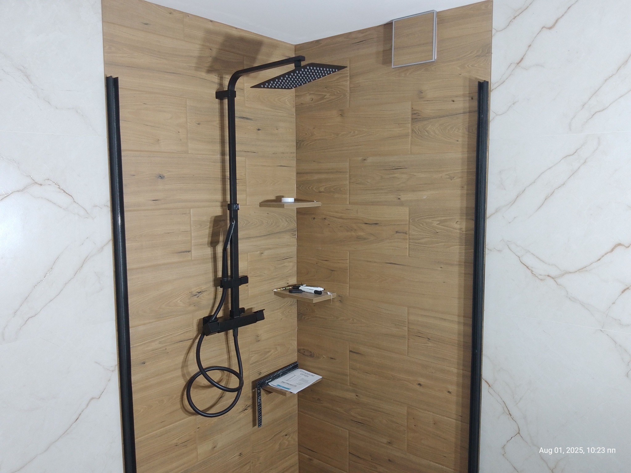 Nowoczesna kabina prysznicowa z czarną armaturą i drewnopodobnymi płytkami. Widoczne półki z akcesoriami i instrukcją montażu. Kontrastujące białe płytki imitujące marmur.