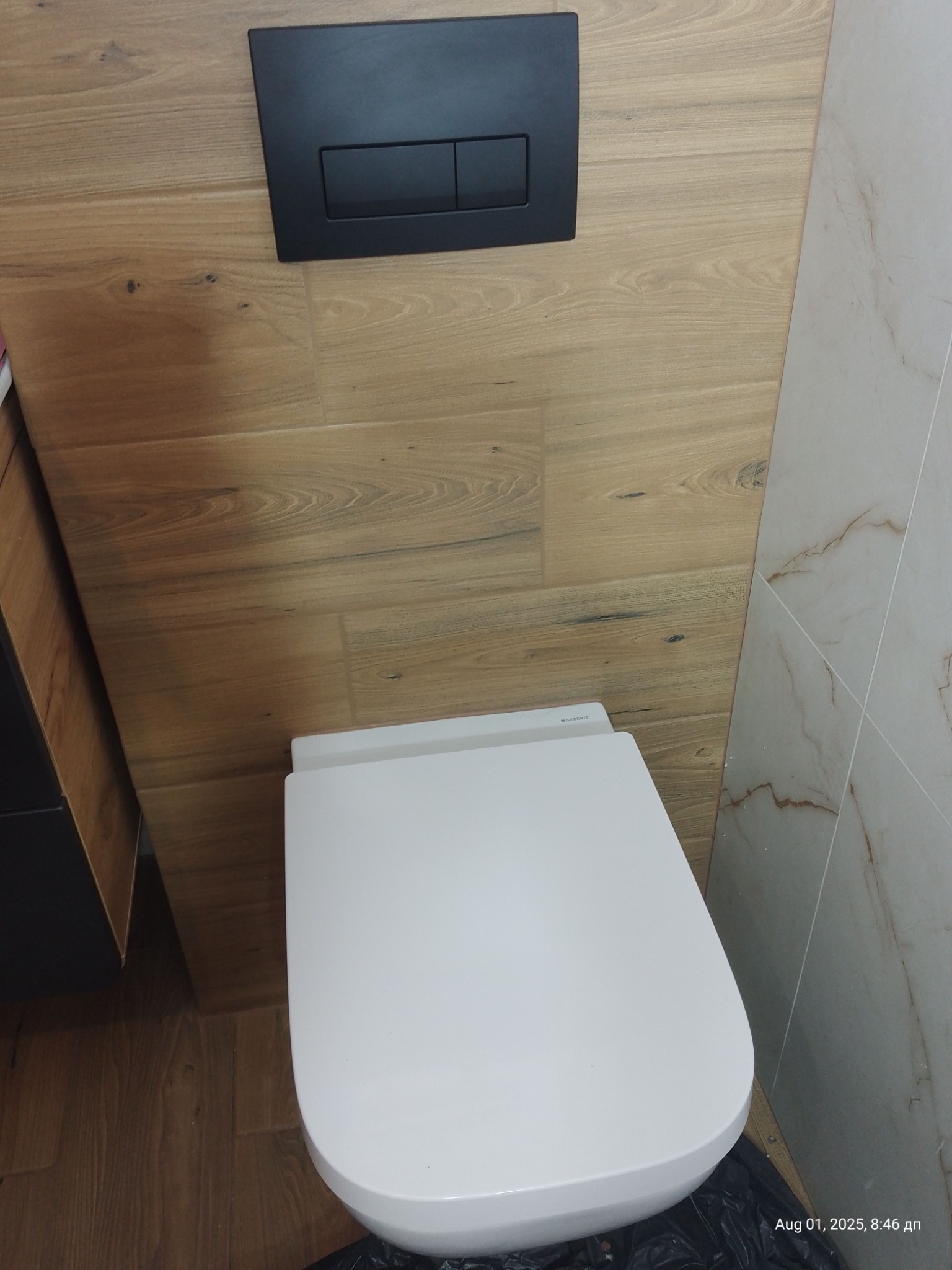 Nowoczesna toaleta podwieszana Geberit z czarnym panelem sterującym na ścianie wyłożonej płytkami imitującymi drewno, sąsiadująca z marmurową ścianą.