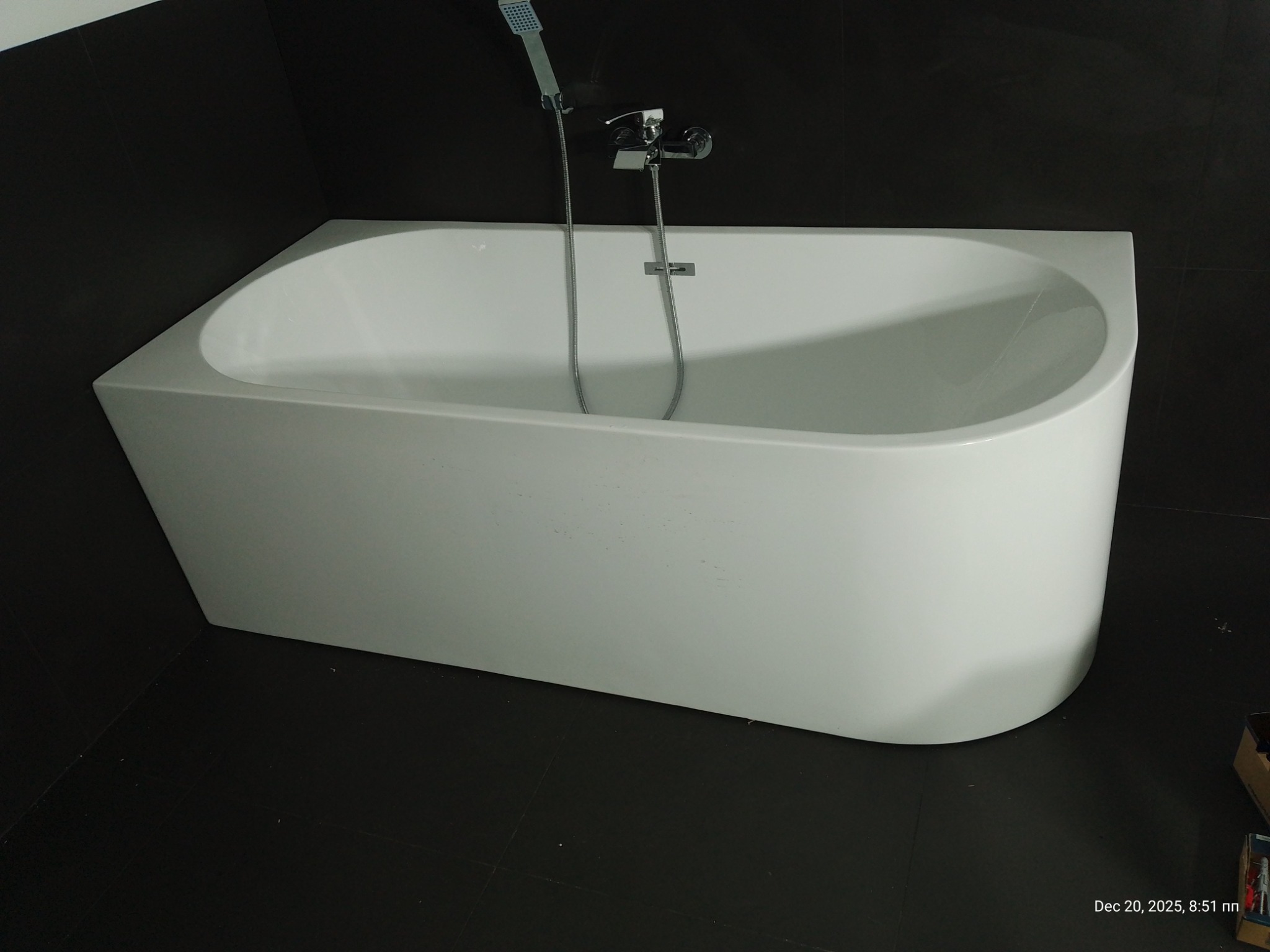 Biała, asymetryczna wanna z chromowaną armaturą na tle czarnych płytek. Minimalistyczny design, nowoczesne wykończenie łazienki. Widok z góry, oświetlenie naturalne.