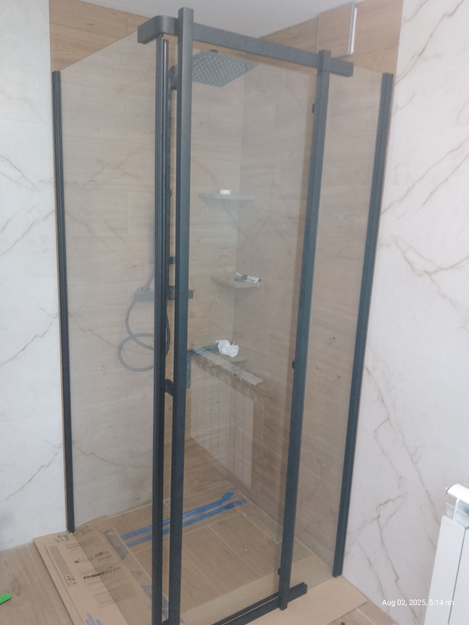 Nowoczesna kabina prysznicowa z czarnymi profilami i deszczownicą, wykończona płytkami imitującymi marmur i drewno. Widoczne zabezpieczenia podłogi.