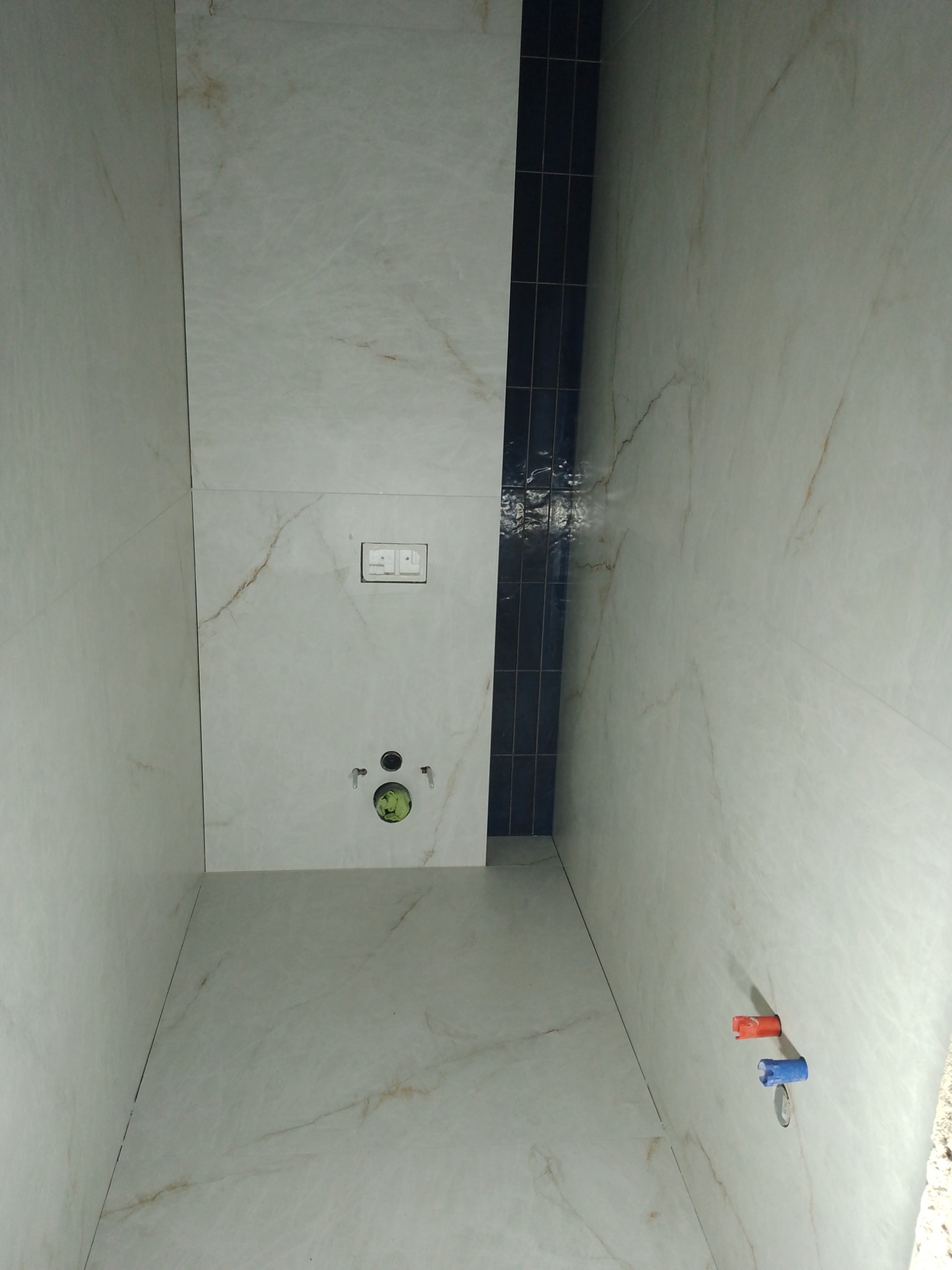 Wykończona łazienka z marmurowymi płytkami, pionowy pas ciemnych płytek, podtynkowy panel WC i wyprowadzenia wody z zaślepkami. Minimalistyczny design.