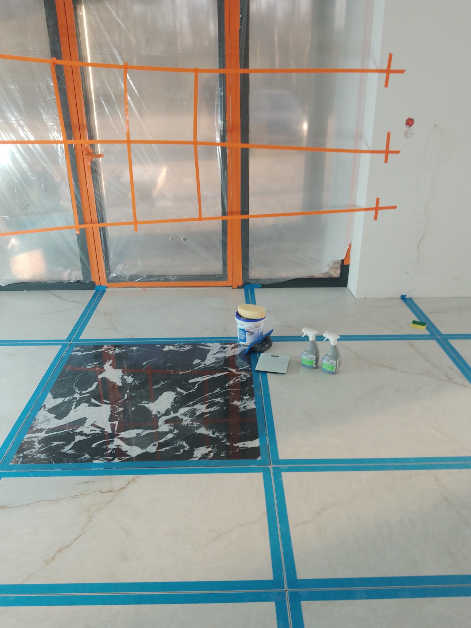 Układanie płytek podłogowych z użyciem taśmy maskującej i narzędzi. Widoczna część ułożonej podłogi z czarnym wzorem. Okno zabezpieczone folią malarską.