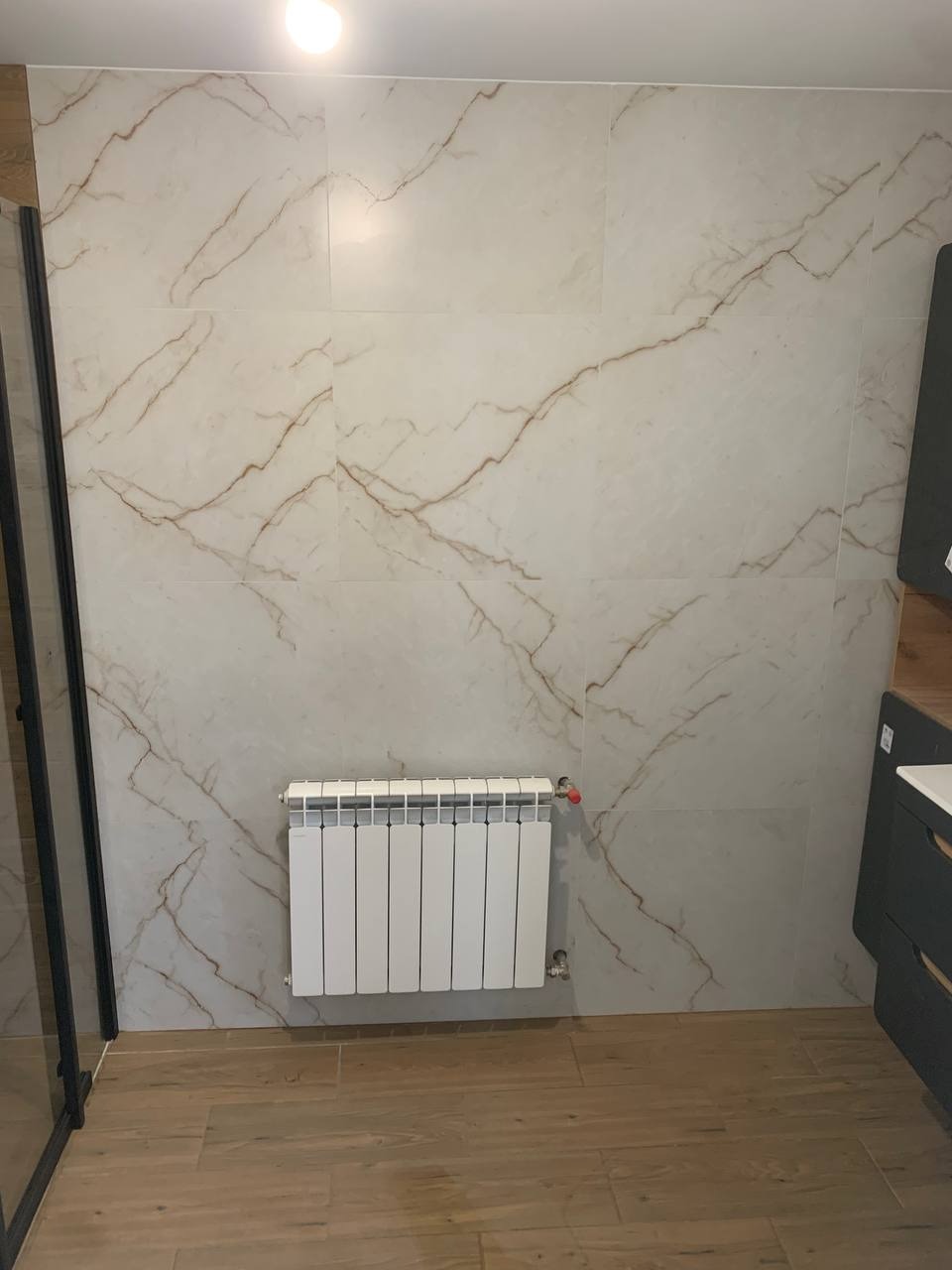 Wykończona łazienka z płytkami imitującymi marmur, białym grzejnikiem i drewnopodobną podłogą. Widoczny fragment kabiny prysznicowej i szafki pod umywalkę.