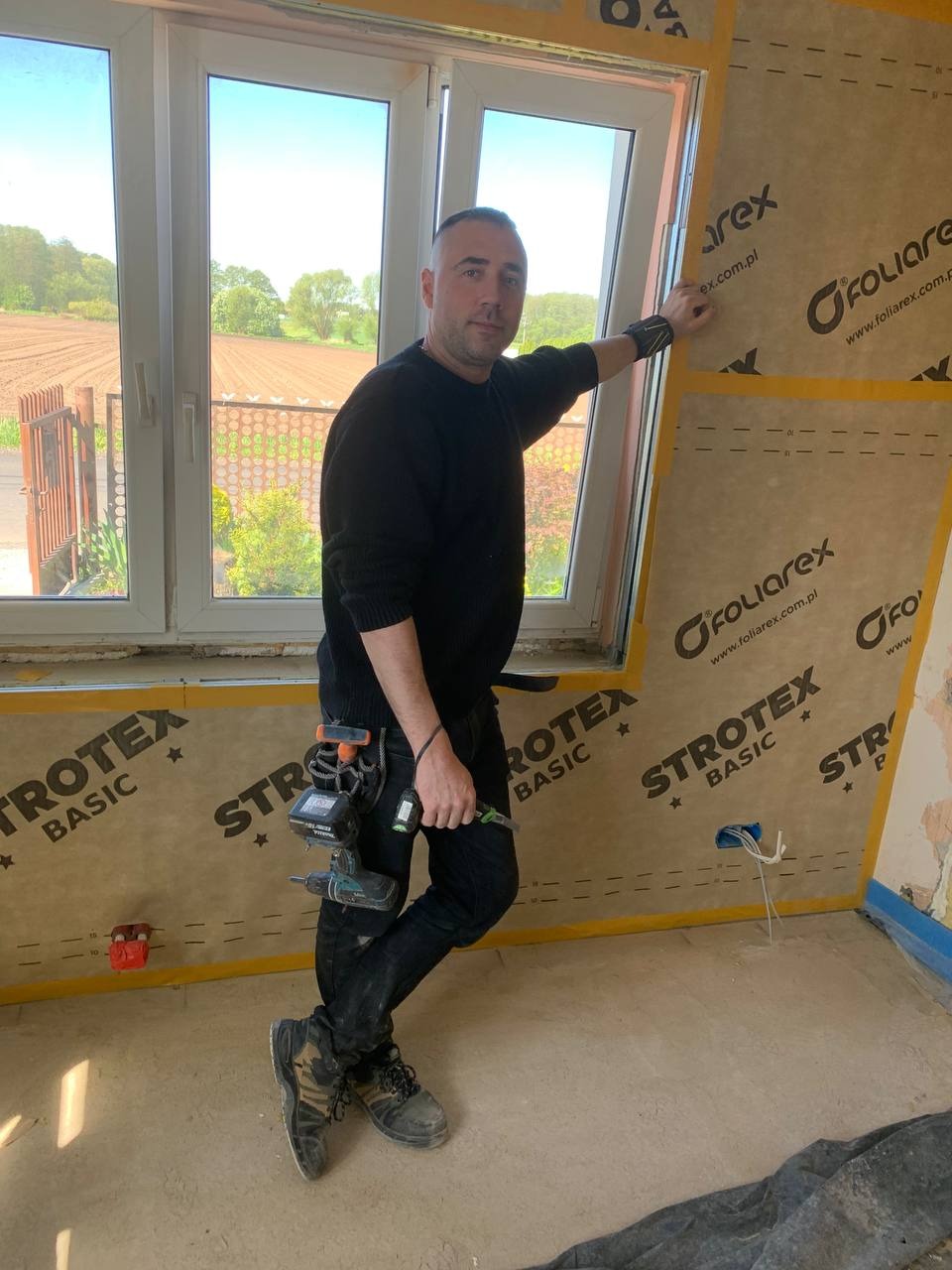 Pracownik z narzędziami montuje izolację termiczną Foliarex na ścianie w nowym budynku. Widoczne okno z widokiem na pole i narzędzia przy pasie.