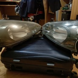 Studio Polerka - Dwie lampy samochodowe, jedna matowa, druga czysta, leżące na czarnej walizce na kółkach. Widoczne tło z półkami i przedmiotami.