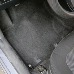 Wnętrze samochodu po praniu tapicerki podłogowej, widoczna czysta, szara wykładzina, fotel kierowcy i fragment deski rozdzielczej.