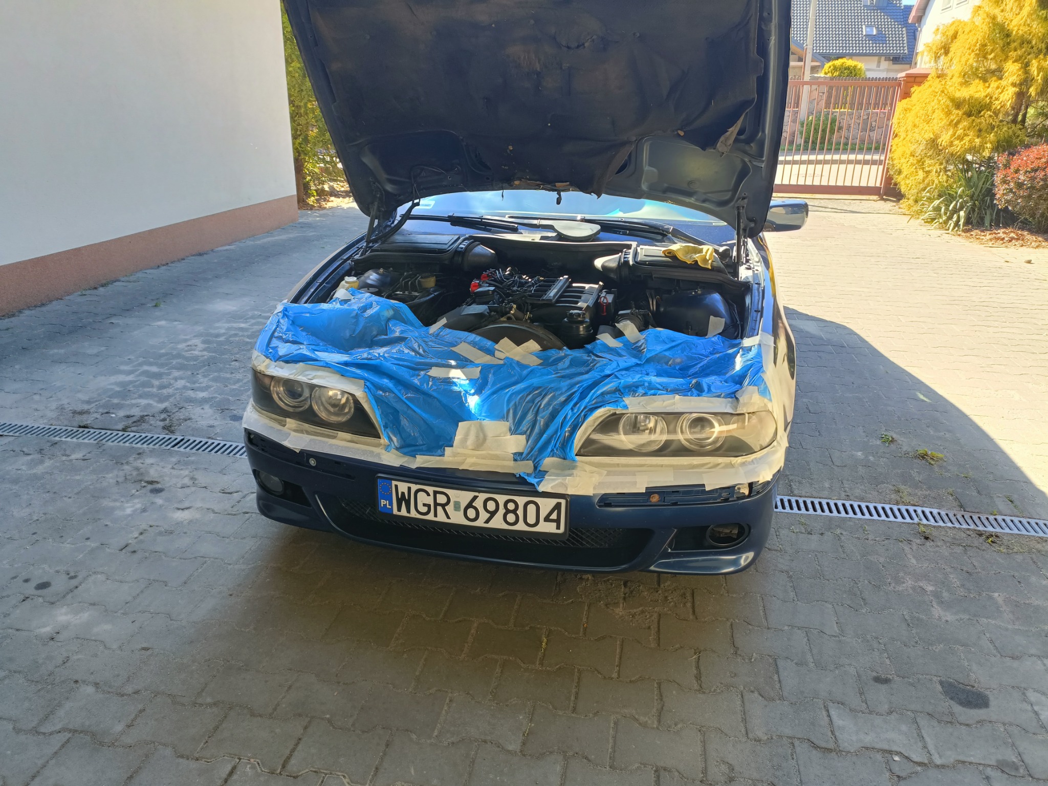 BMW E39 z otwartą maską, przygotowywane do regeneracji lamp. Zabezpieczone folią i taśmą, widoczny silnik. Tablica rejestracyjna WGR 69804.