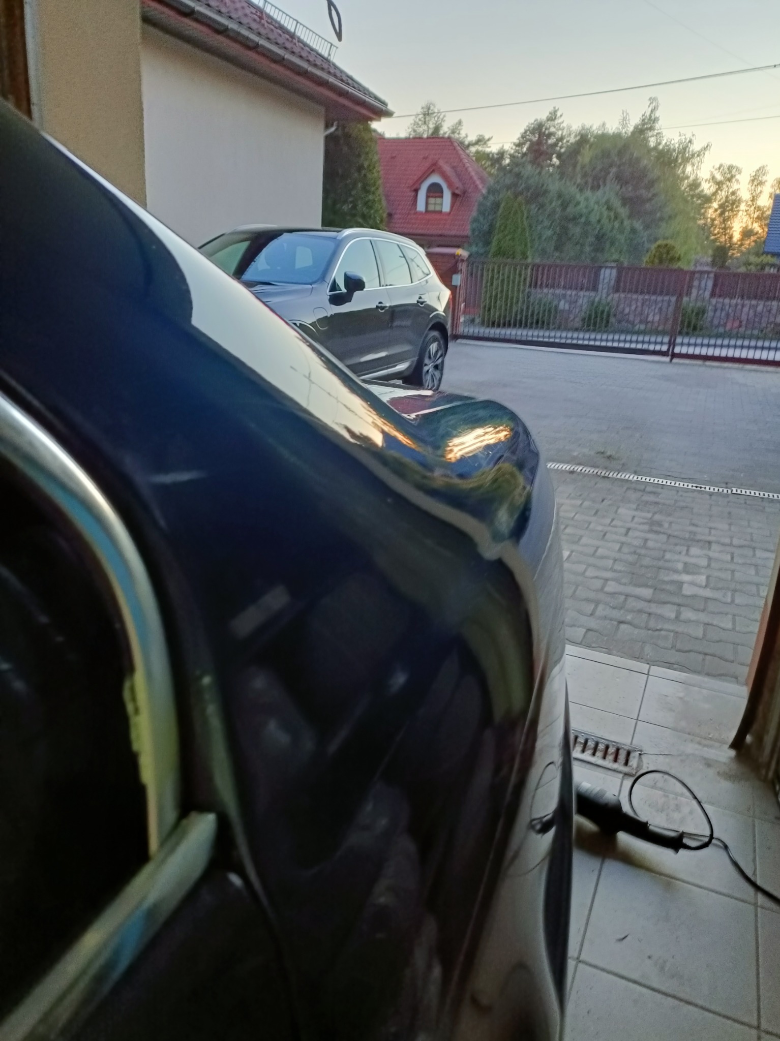 Błyszczący fragment karoserii auta po polerowaniu, z widocznym urządzeniem do polerowania na posadzce. W tle drugi samochód i dom z czerwoną dachówką.