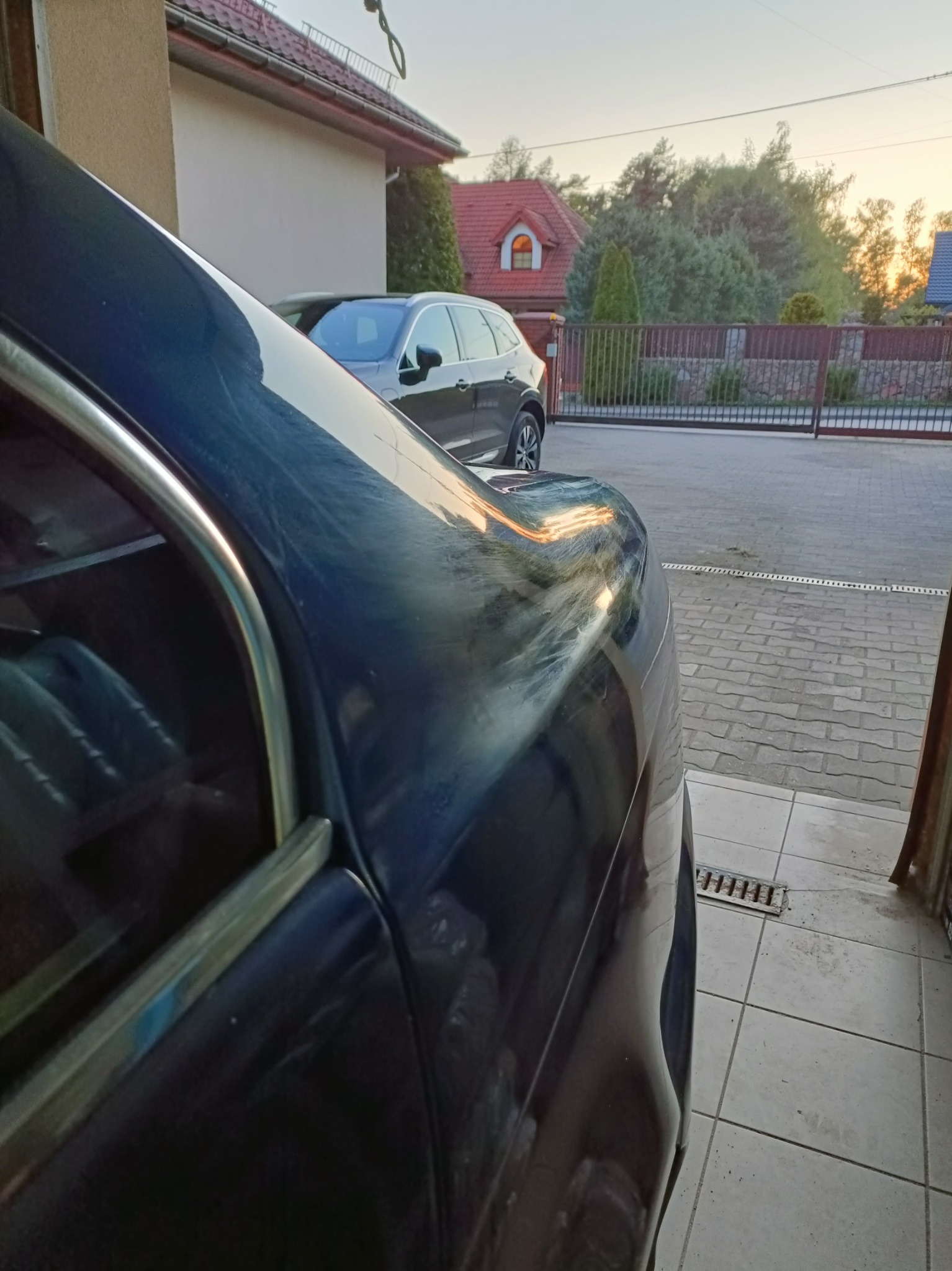Fragment karoserii ciemnego auta z widocznymi smugami i zabrudzeniami, zaparkowanego na zewnątrz, w tle fragment domu i brama wjazdowa.